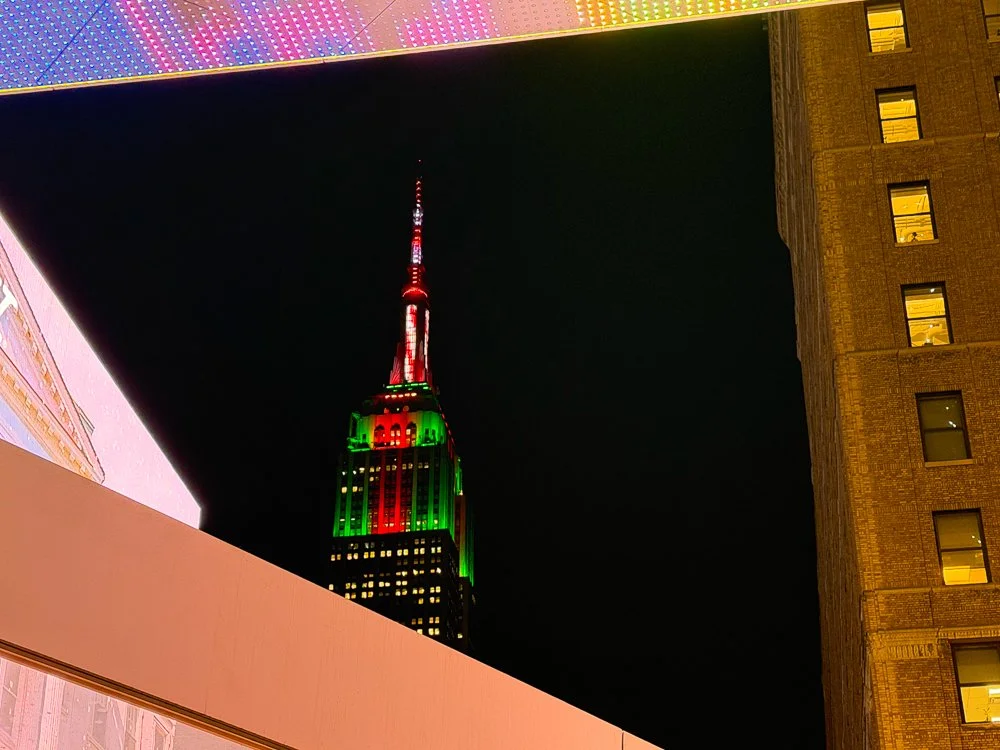 The Empire State Building lit up in red, green, and white lights for Christmas, seen through a gap between two buildings at night.