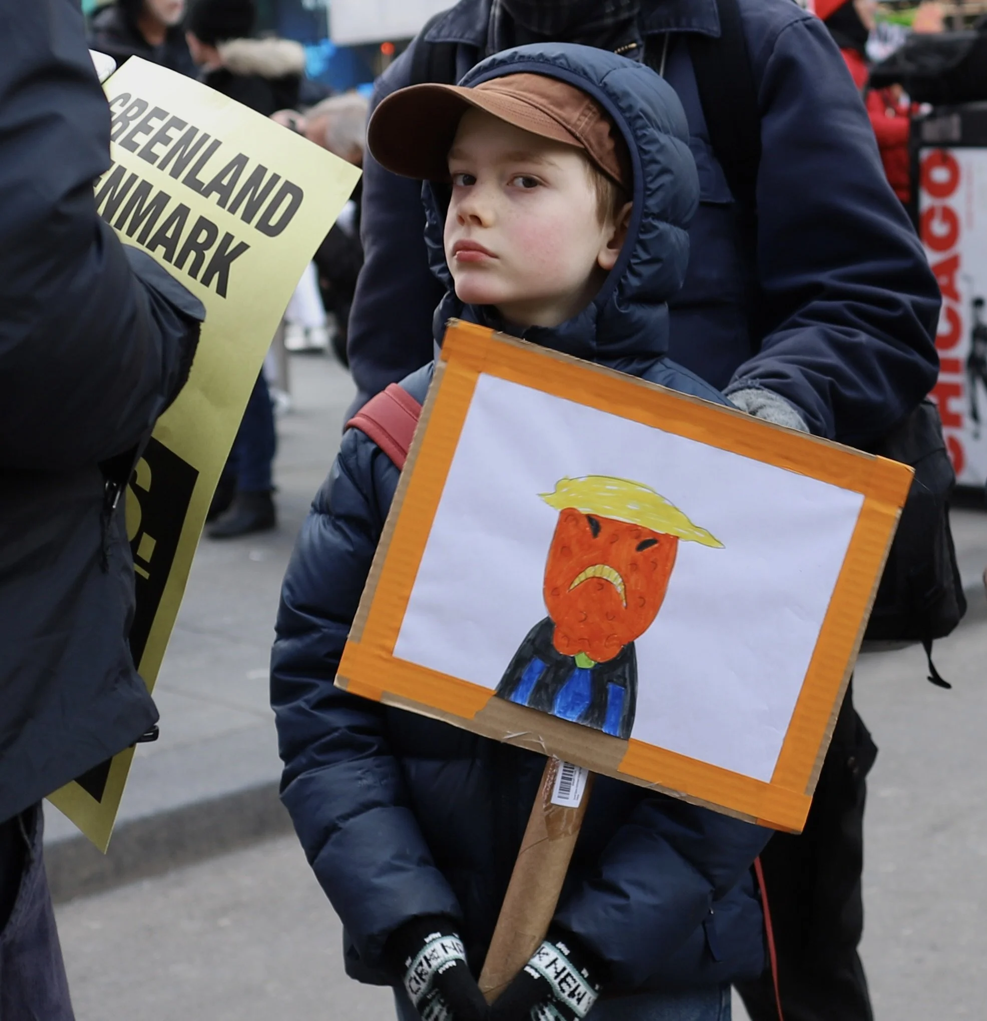 Young boy at a protest holding a sign with a sad face and styled to resemble Donald Trump with blonde hair. The boy is wearing a blue jacket, brown cap, and black gloves. Several other protest signs are visible in the background.