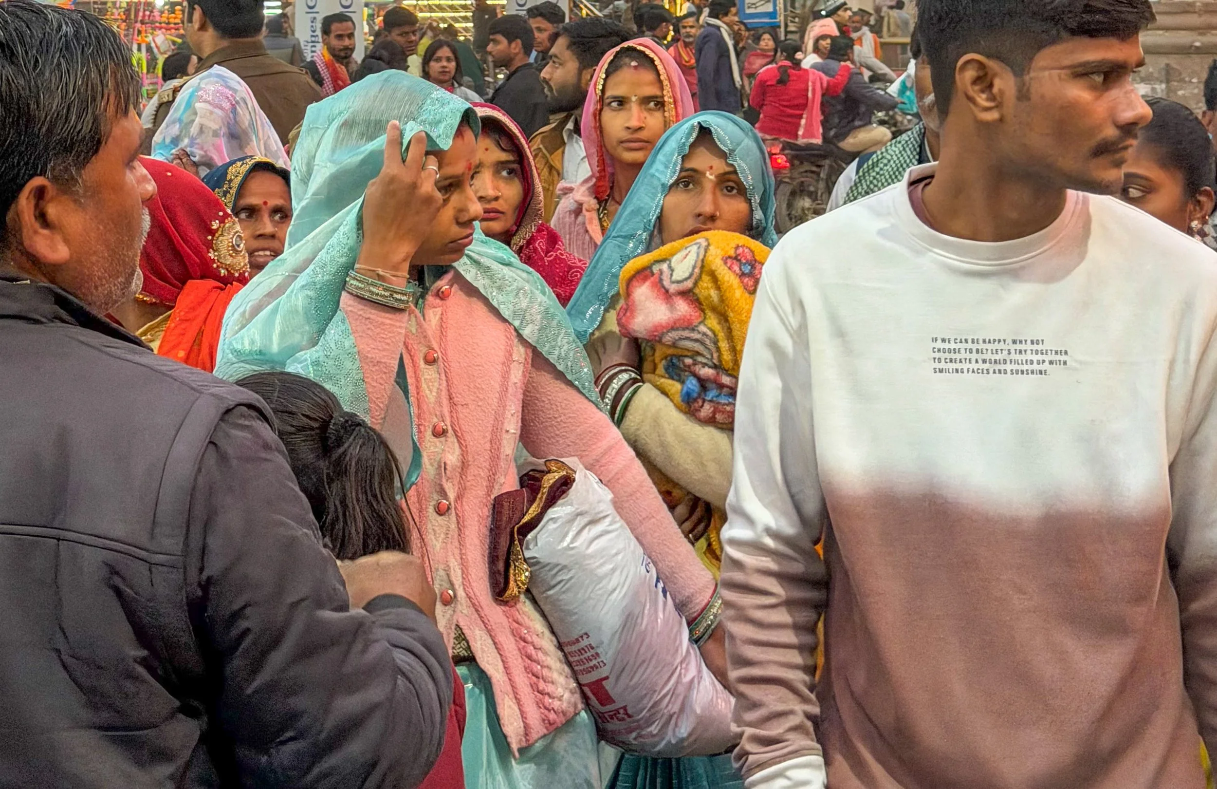 A crowd of people in a busy outdoor setting, including women in colorful traditional Indian attire with scarves and men in casual clothing. Some individuals appear to be waiting or observing, with a woman in the center adjusting her headscarf and ano