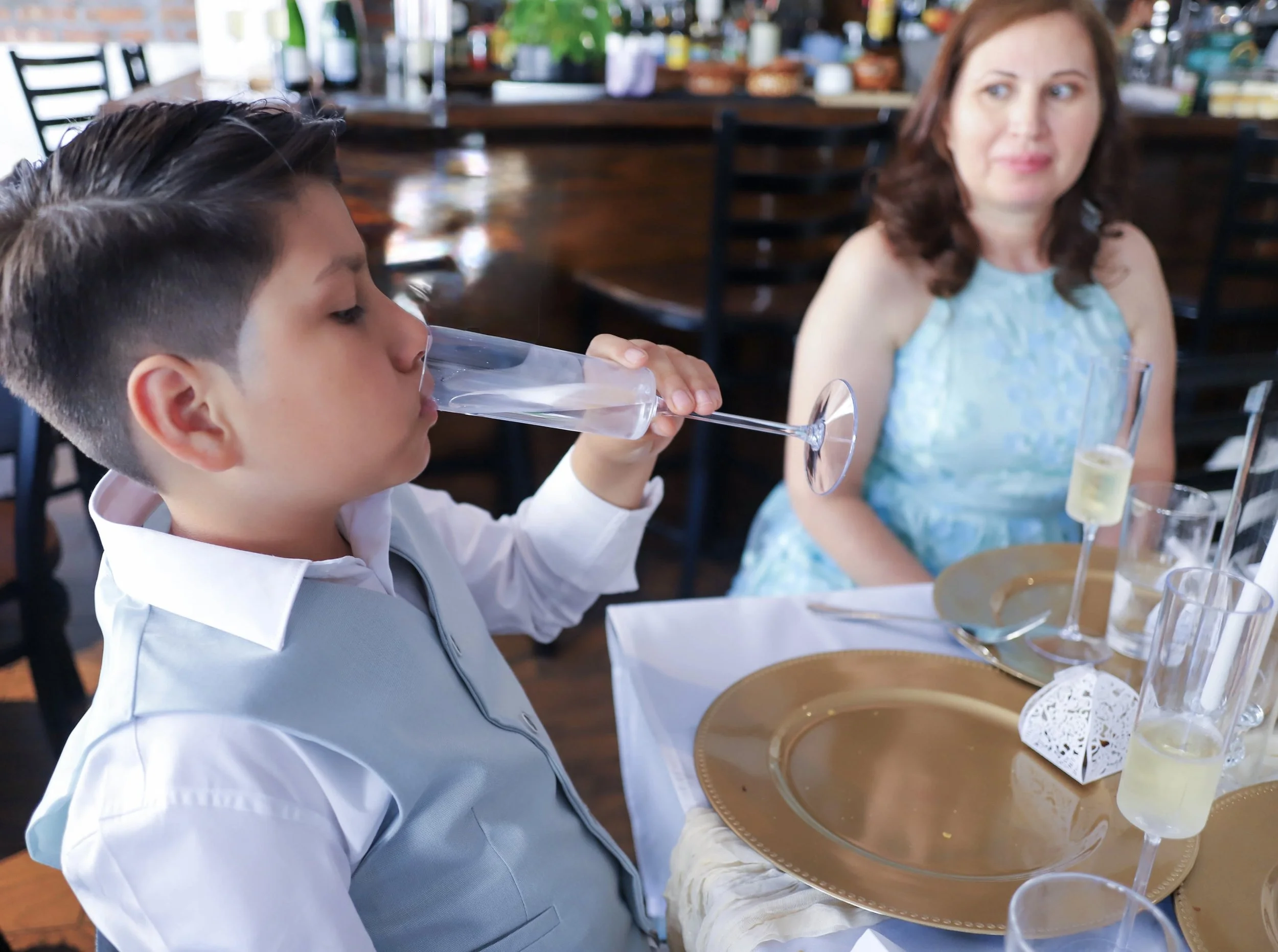 A young boy in a white shirt and light blue vest drinking from a champagne flute at a formal table setting, with a woman in a blue dress sitting nearby in a restaurant or event venue.