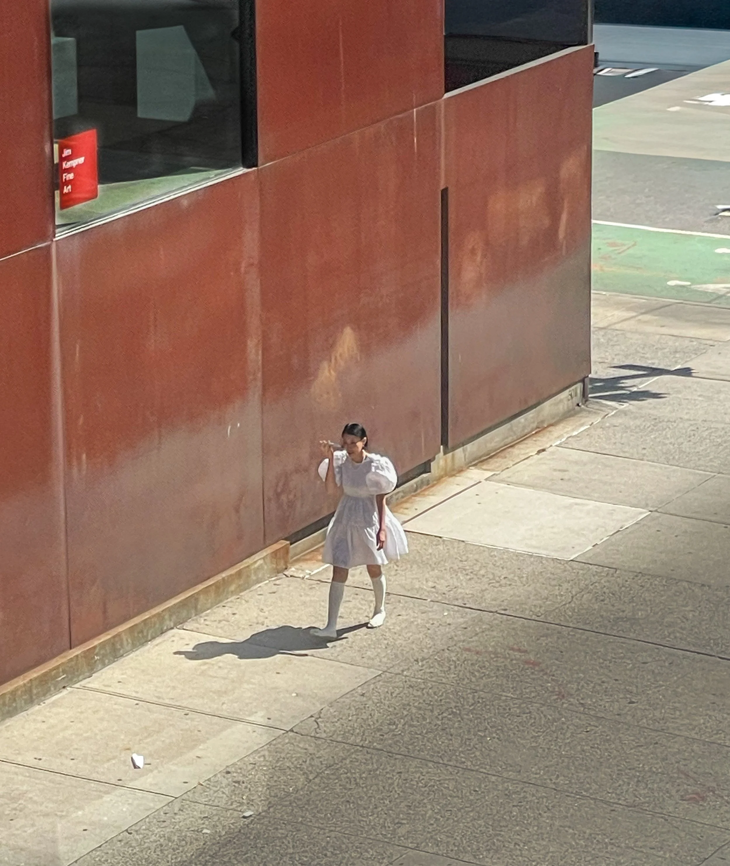 A girl in a white dress and knee-high socks walking on a sidewalk next to a large rust-colored building, carrying a stick over her shoulder.