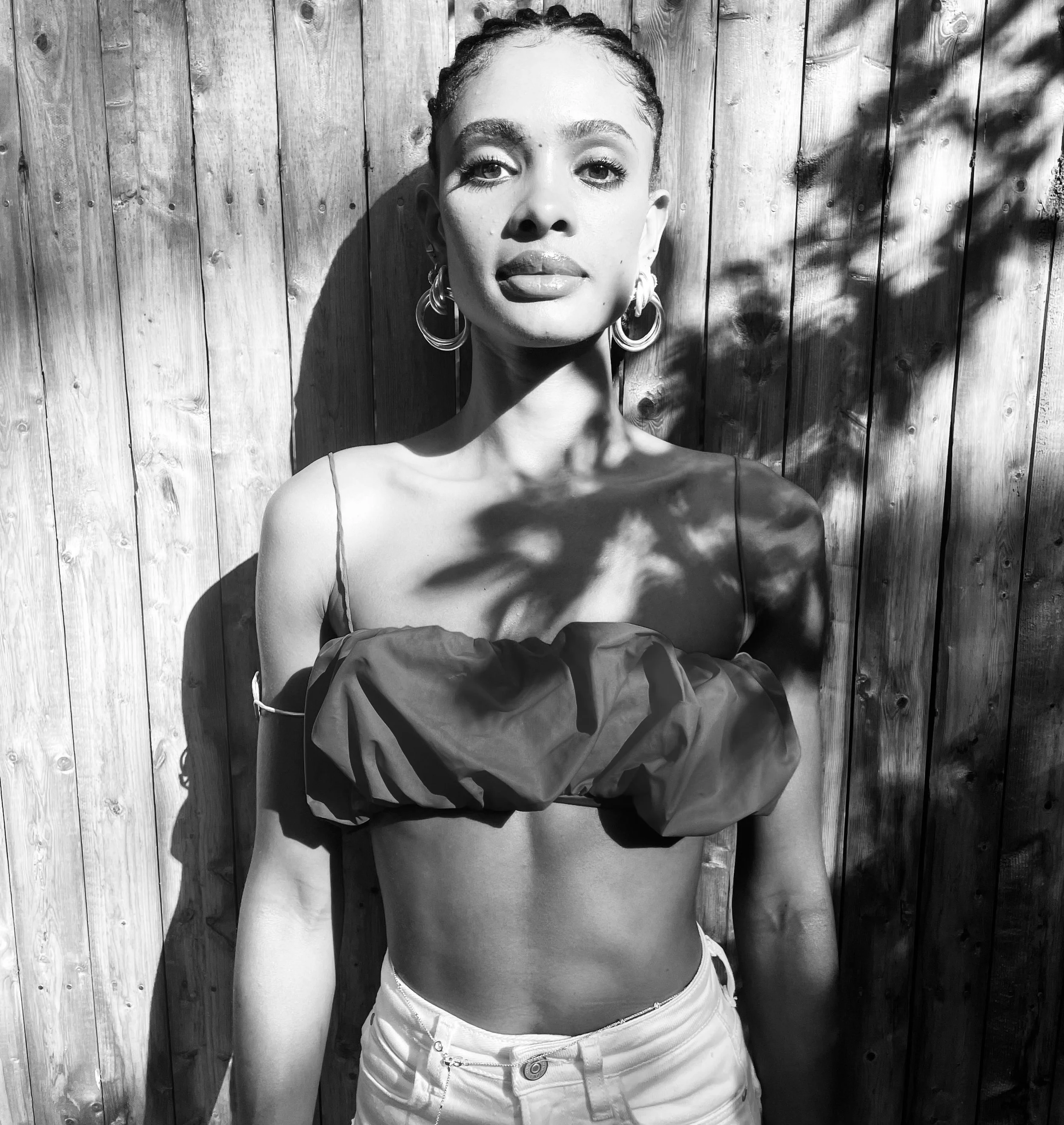 A woman with braided hair and hoop earrings standing in front of a wooden fence, casting shadows from nearby leaves, looking at the camera with her tongue slightly out.
