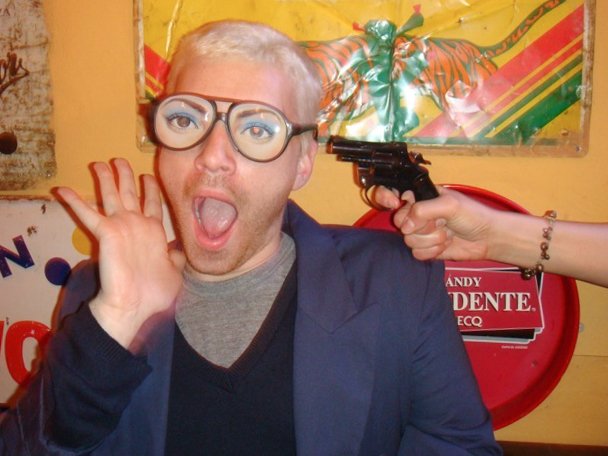 A man with short blonde hair, glasses, and wearing a blue blazer and gray shirt, reacts with surprise as a hand points a gun at his head. The background includes a colorful carnival-style decoration, a red mailbox, and a sign referencing a candidate.