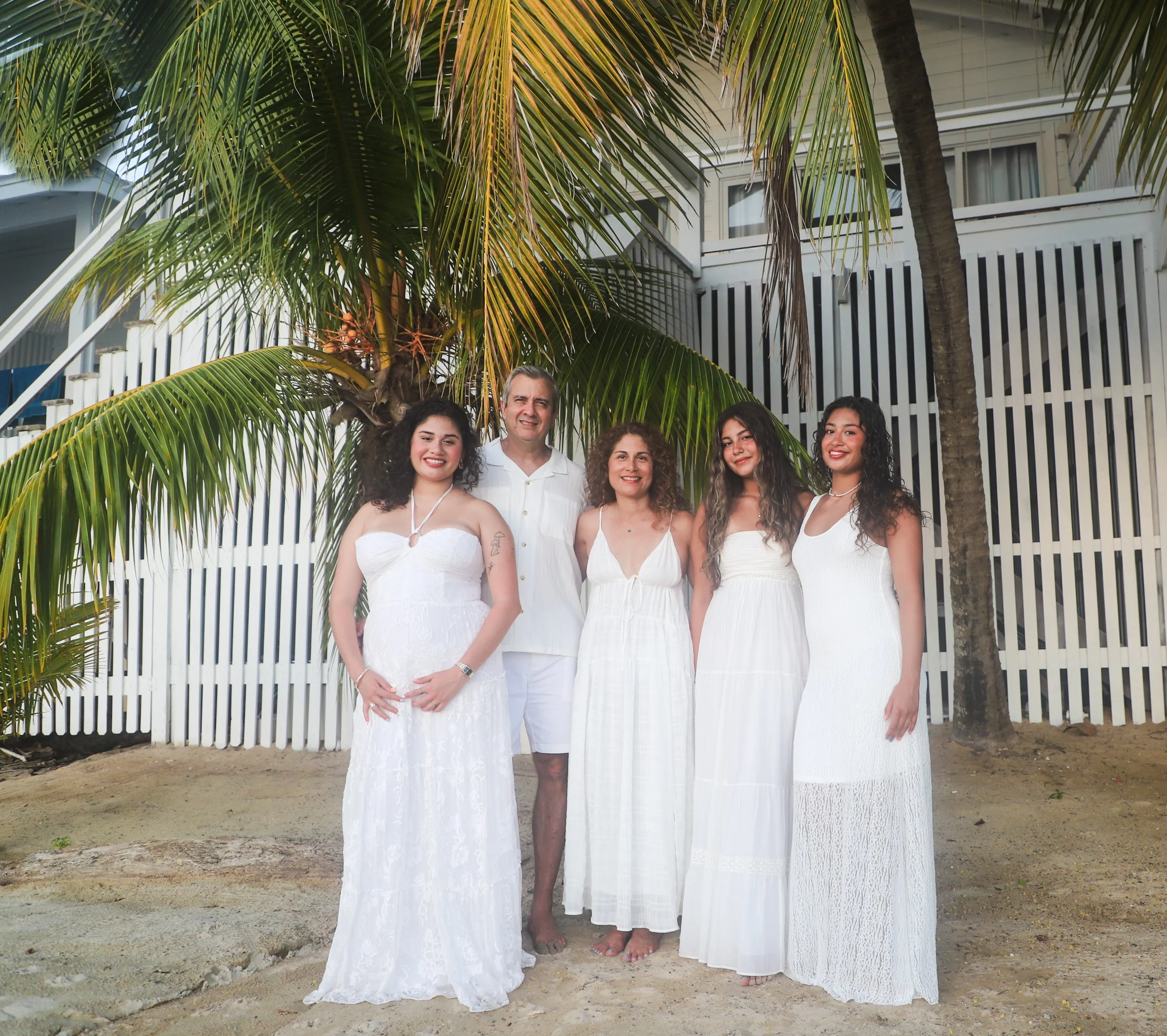 A group of five people standing on a beach in front of a white picket fence and palm trees, smiling at the camera. The group consists of four women dressed in white dresses and a man in white shorts and shirt.