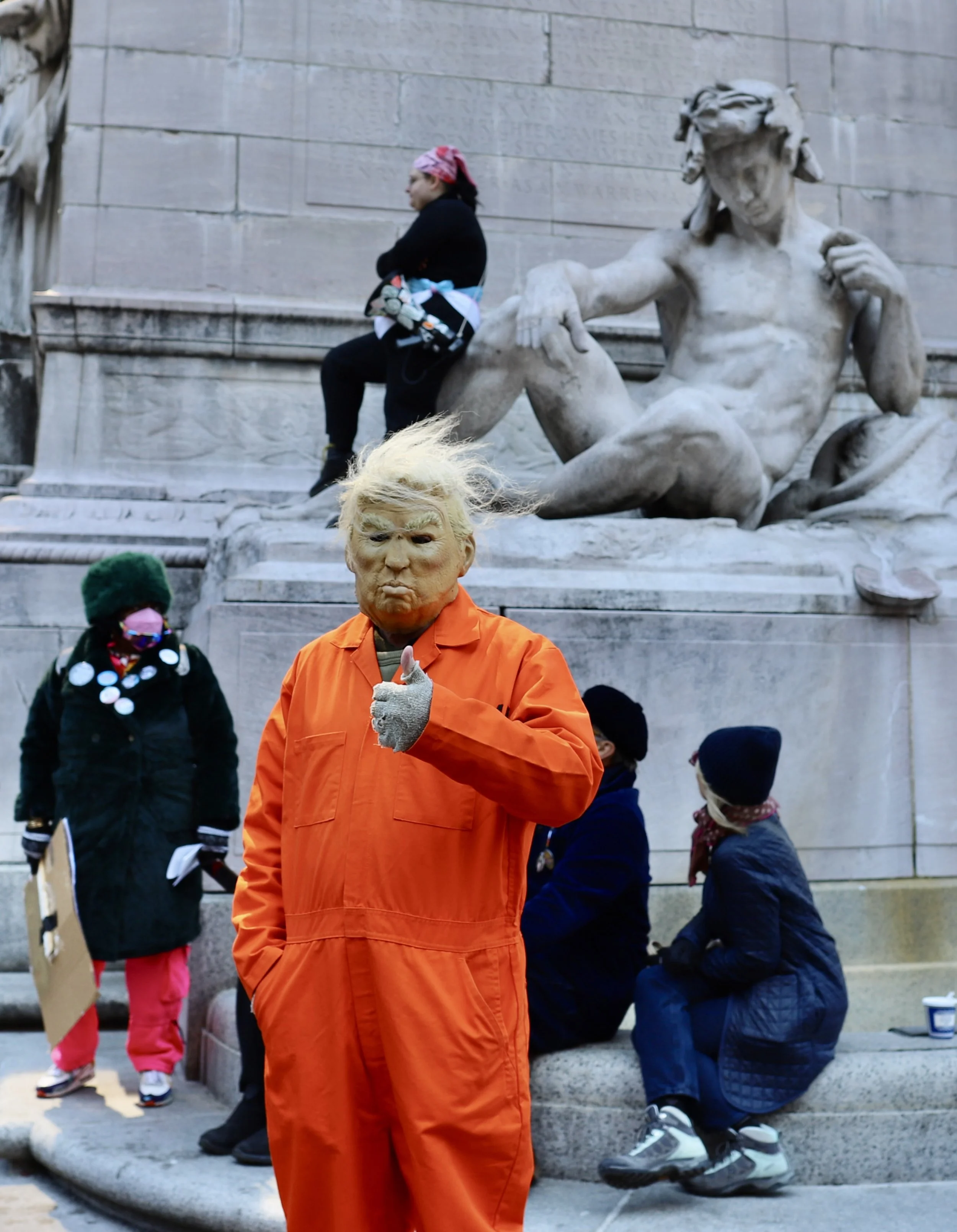 Person dressed as Donald Trump in an orange prison jumpsuit giving a thumbs-up in front of a crowd, with people sitting and standing near the Lincoln Memorial.