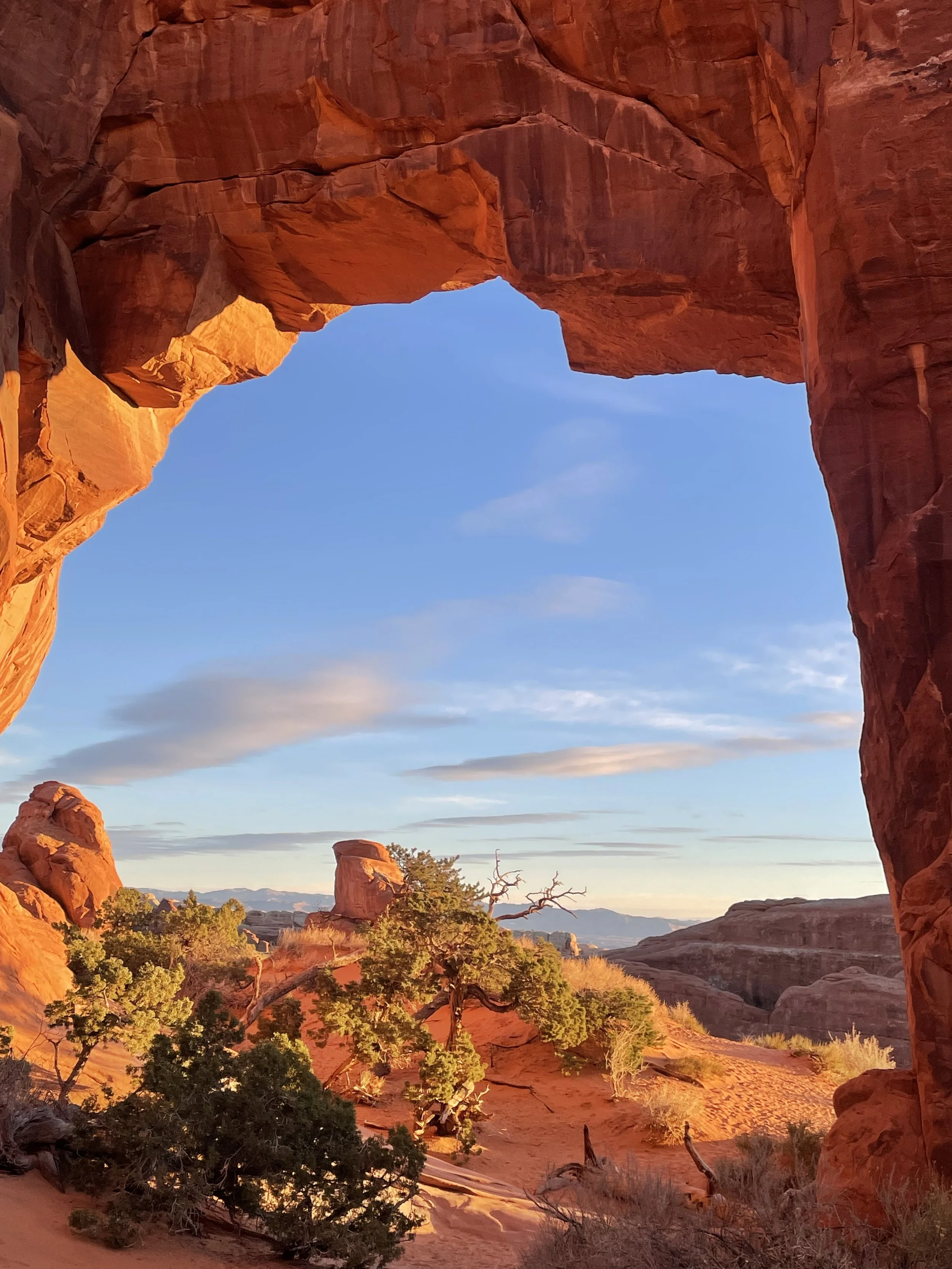 View through a red rock arch at sunset or sunrise, showing desert vegetation and distant mountains under a partly cloudy sky.