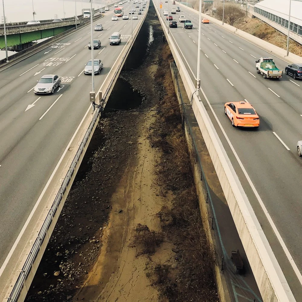 Aerial view of a highway with two lanes of traffic in each direction separated by a divider. Vehicles are traveling in both directions. The right side of the highway has a concrete barrier, and the left side has a green bridge structure over the road
