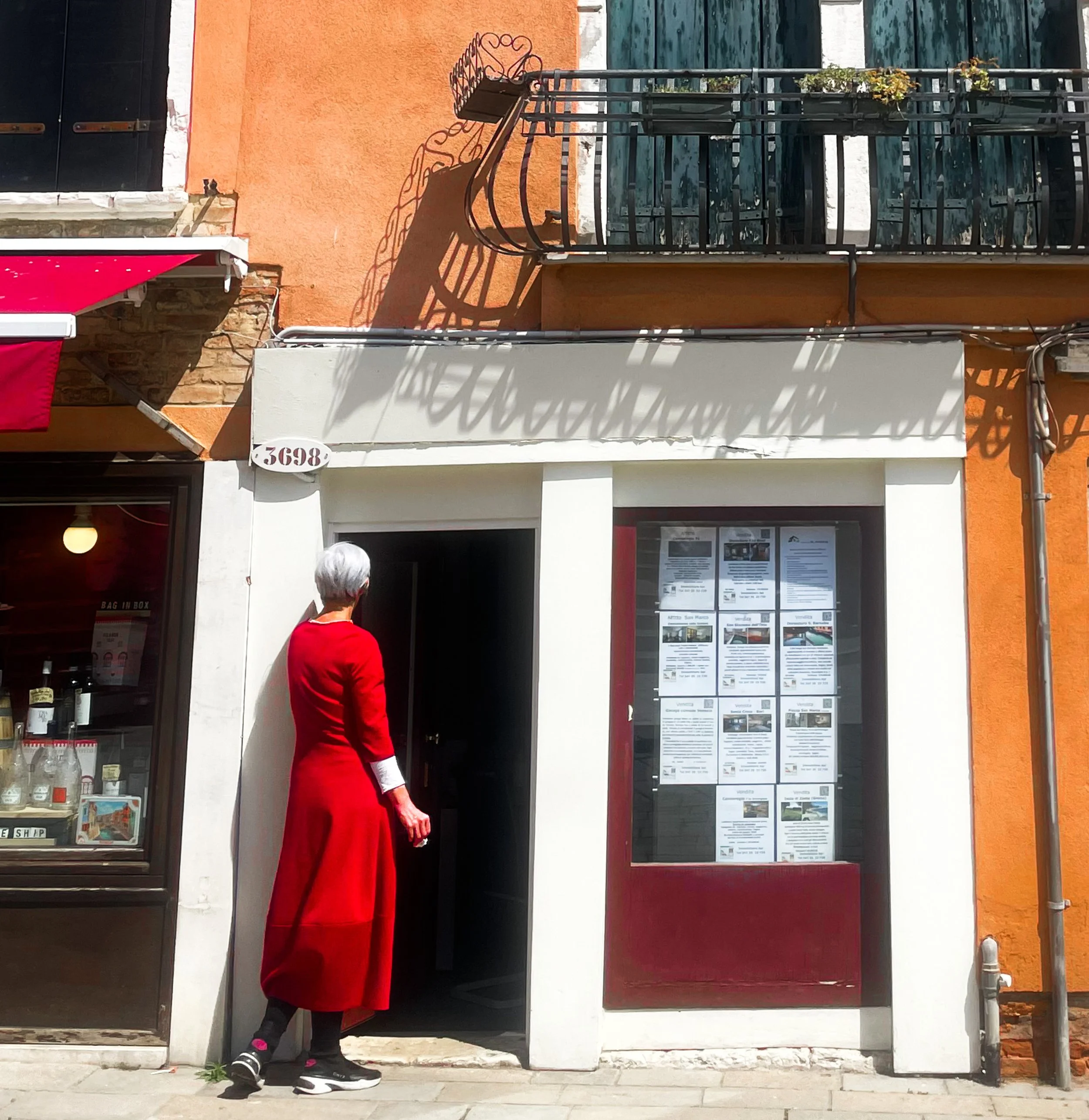 A woman in a red dress and sneakers stands outside a restaurant, looking at the menu posted on the window. The building has an orange and white facade with a balcony above.