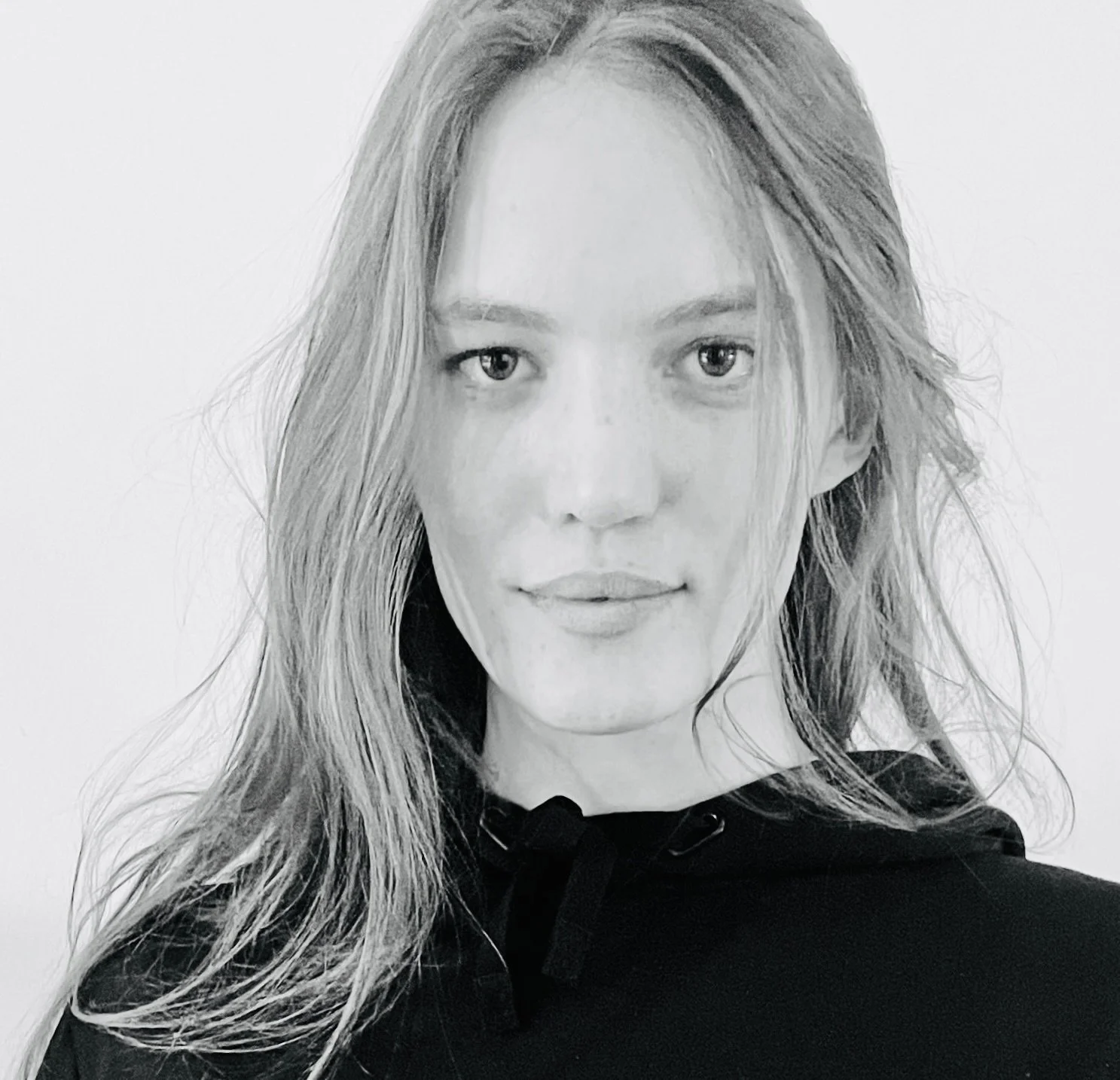 A black and white photo of a young woman with long hair, wearing a hoodie, looking directly at the camera with a slight smile.