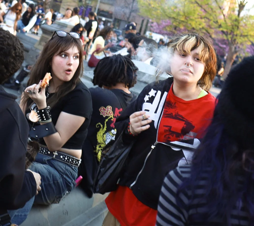 Young people socializing outdoors, with two individuals in the foreground smoking and eating pizza, surrounded by others at a lively gathering with trees and people in the background.