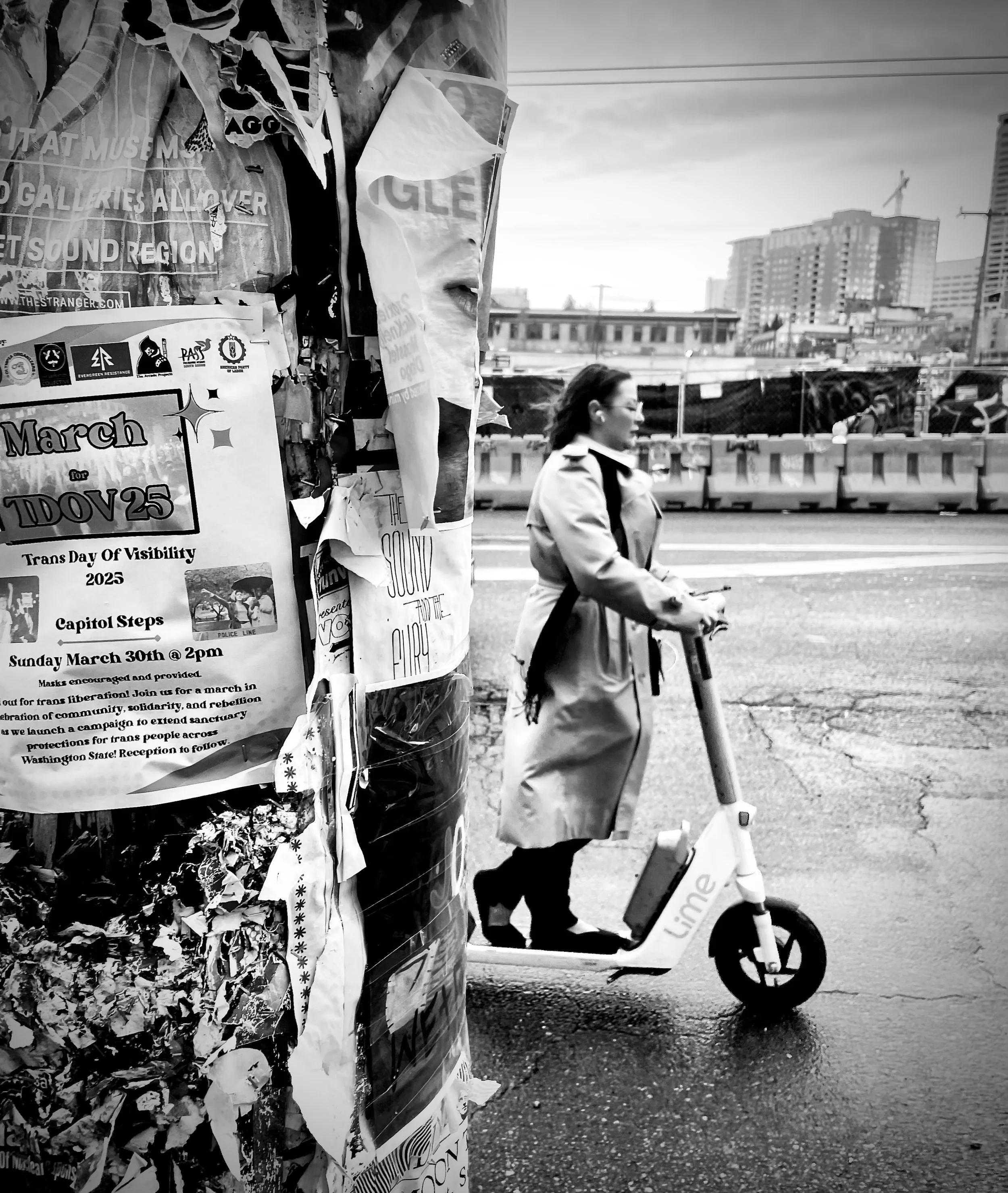 A woman riding an electric scooter on a city street near a cluttered pole covered with posters, with tall buildings and construction cranes in the background.