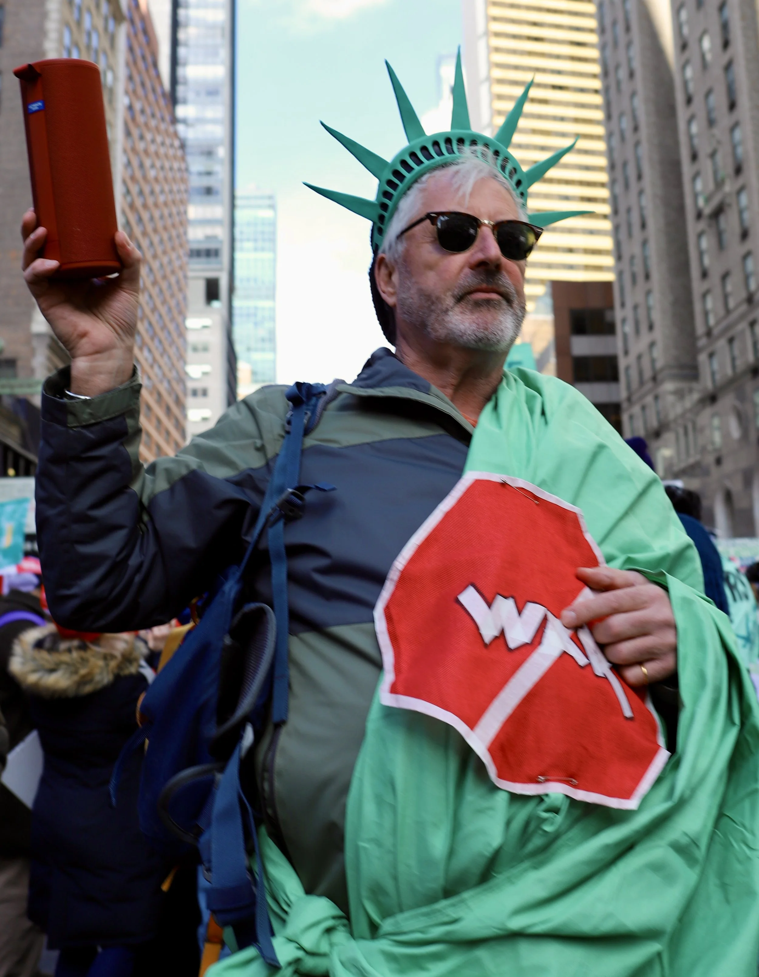 A man dressed as the Statue of Liberty holding a red stop sign during a protest or demonstration in an urban setting.
