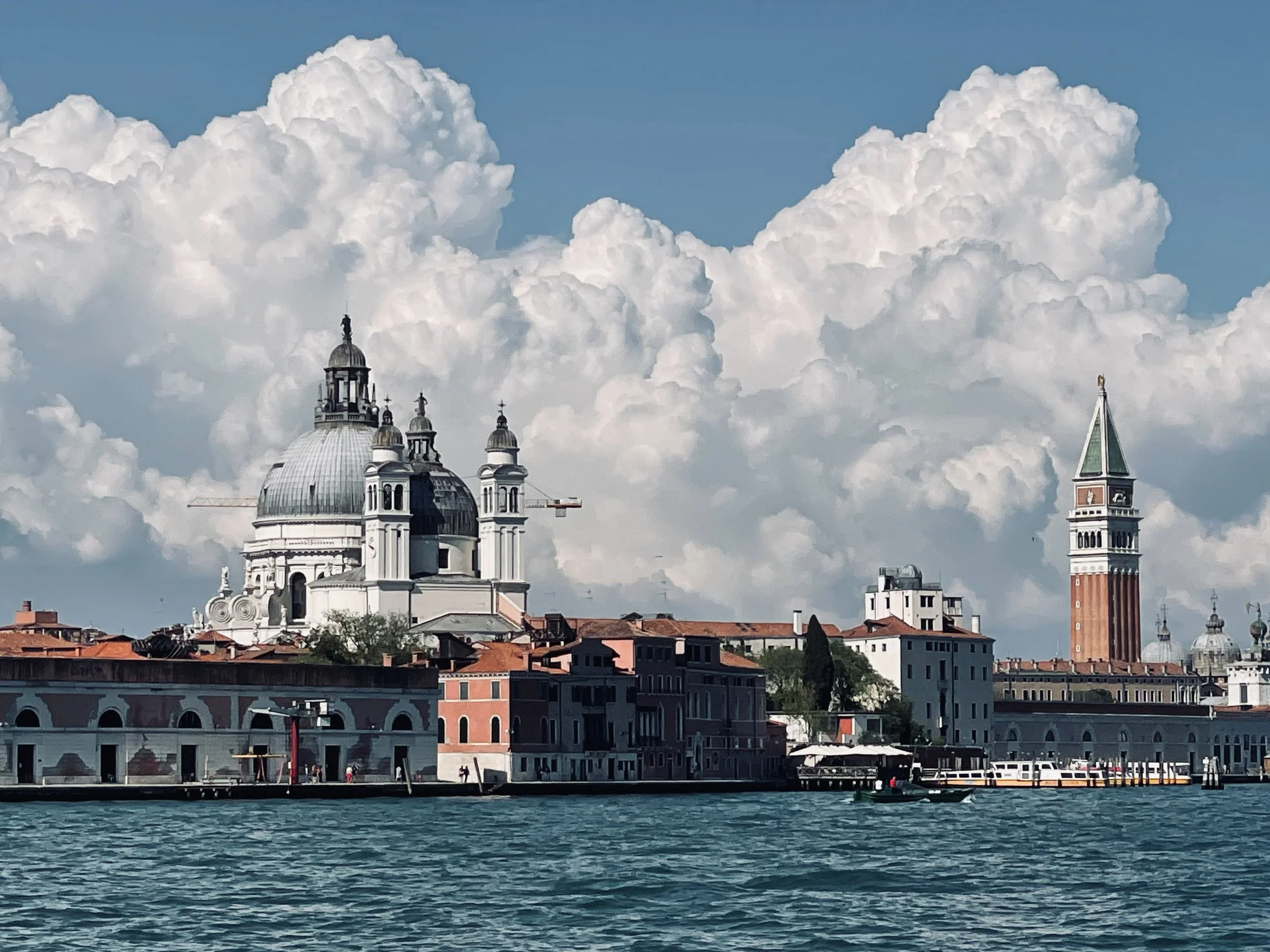 Waterfront view of Venice, Italy, featuring a large baroque-style church with a prominent dome and smaller towers, and a tall bell tower with a pointed roof, under a sky filled with white clouds.