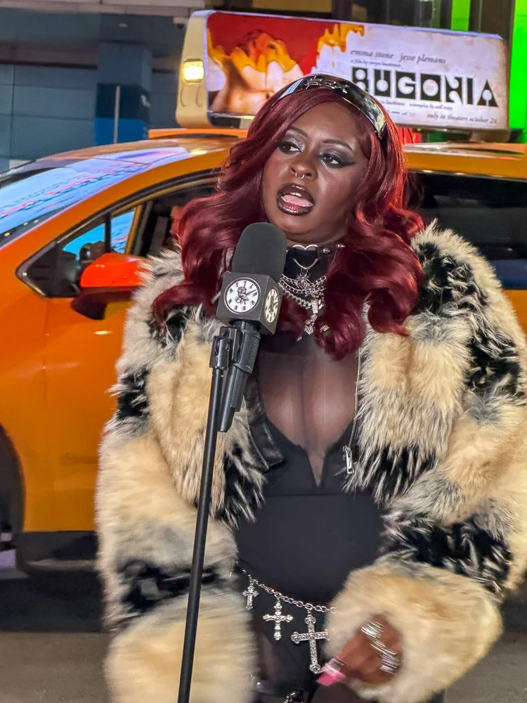 A woman with red hair speaking into a microphone on a city street at night, wearing a fur coat, dark makeup, and jewelry, with a yellow taxi and an illuminated billboard in the background.