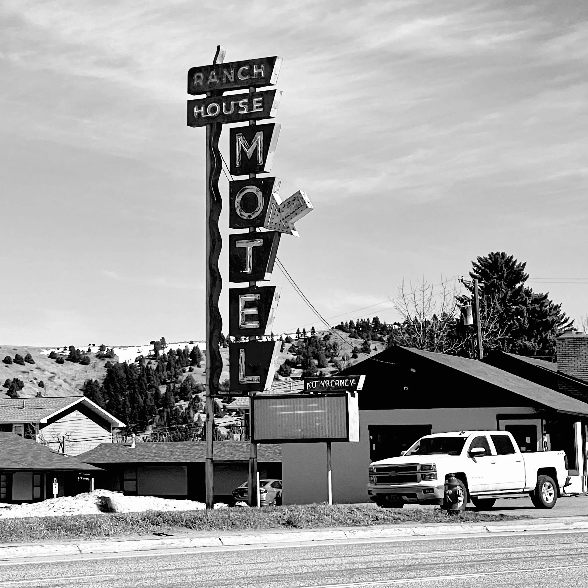 A vintage vertical motel sign reading 'Ranch House Motel' with an arrow pointing to the right, next to a small building with a white pickup truck parked in front, on a street with a hillside covered in trees in the background.
