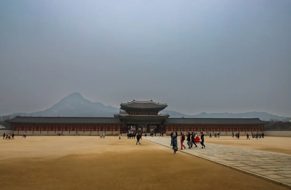 Traditional Asian palace with a central pagoda-style roof, surrounded by a long wall, with visitors walking towards it on a wide plaza under an overcast sky, with mountains in the background.