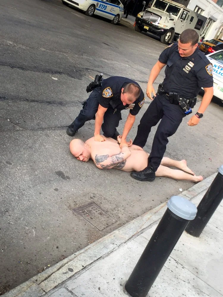 Two police officers are kneeling on a city street, detaining a naked man lying on the ground. The officers are holding the man down by his arms. Police cars are visible parked in the background.