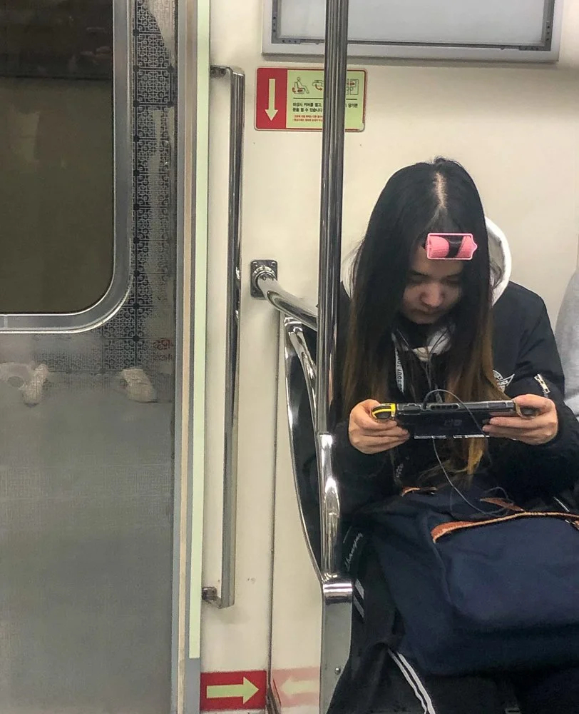 A woman with long black hair is sitting on a train seat, looking at her phone, with a pink roller in her hair. She is wearing a black jacket and has a backpack on her lap.