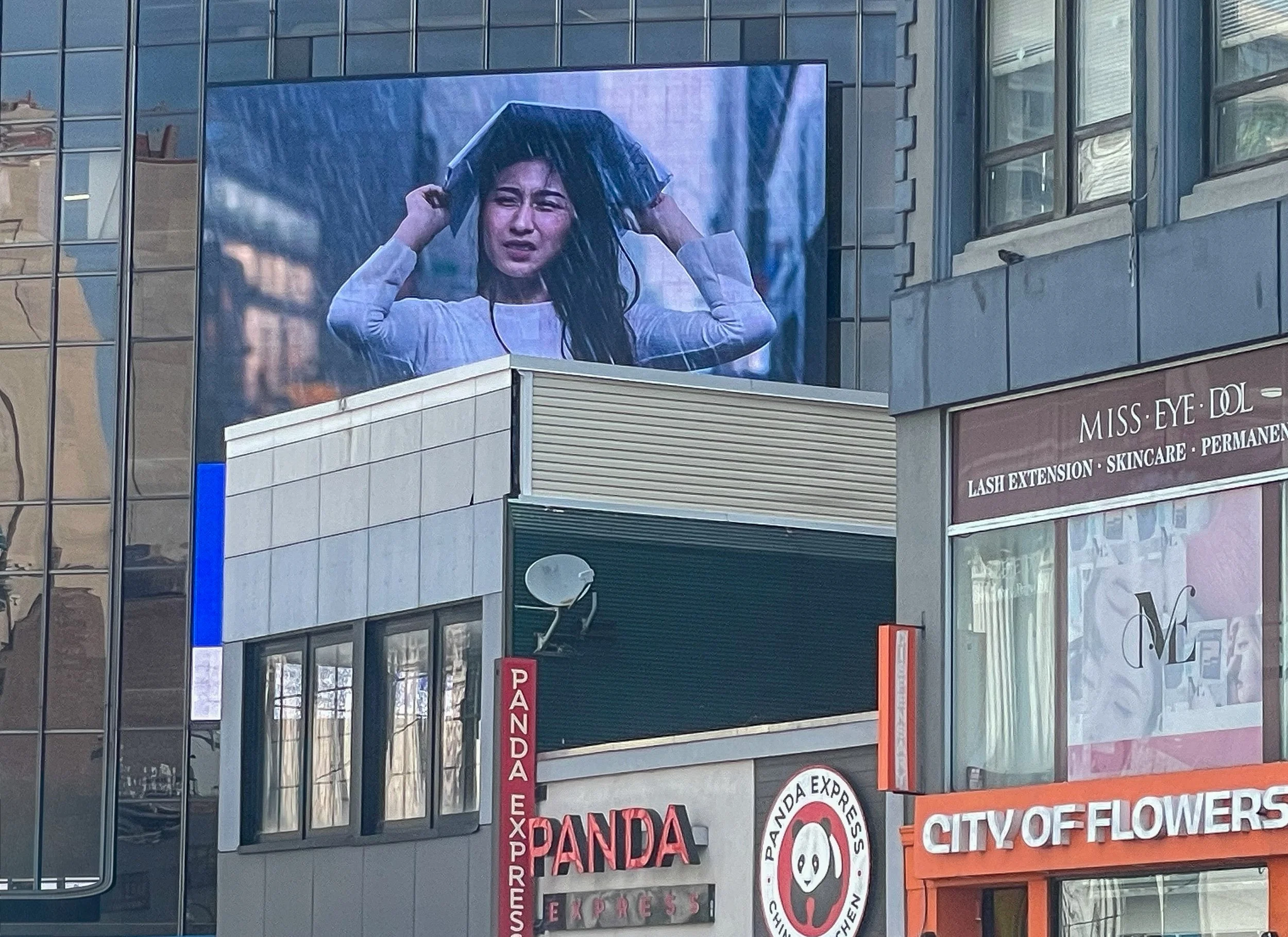 A large outdoor digital billboard showing a woman with wet hair holding an umbrella against the rain, with buildings and storefronts visible below.