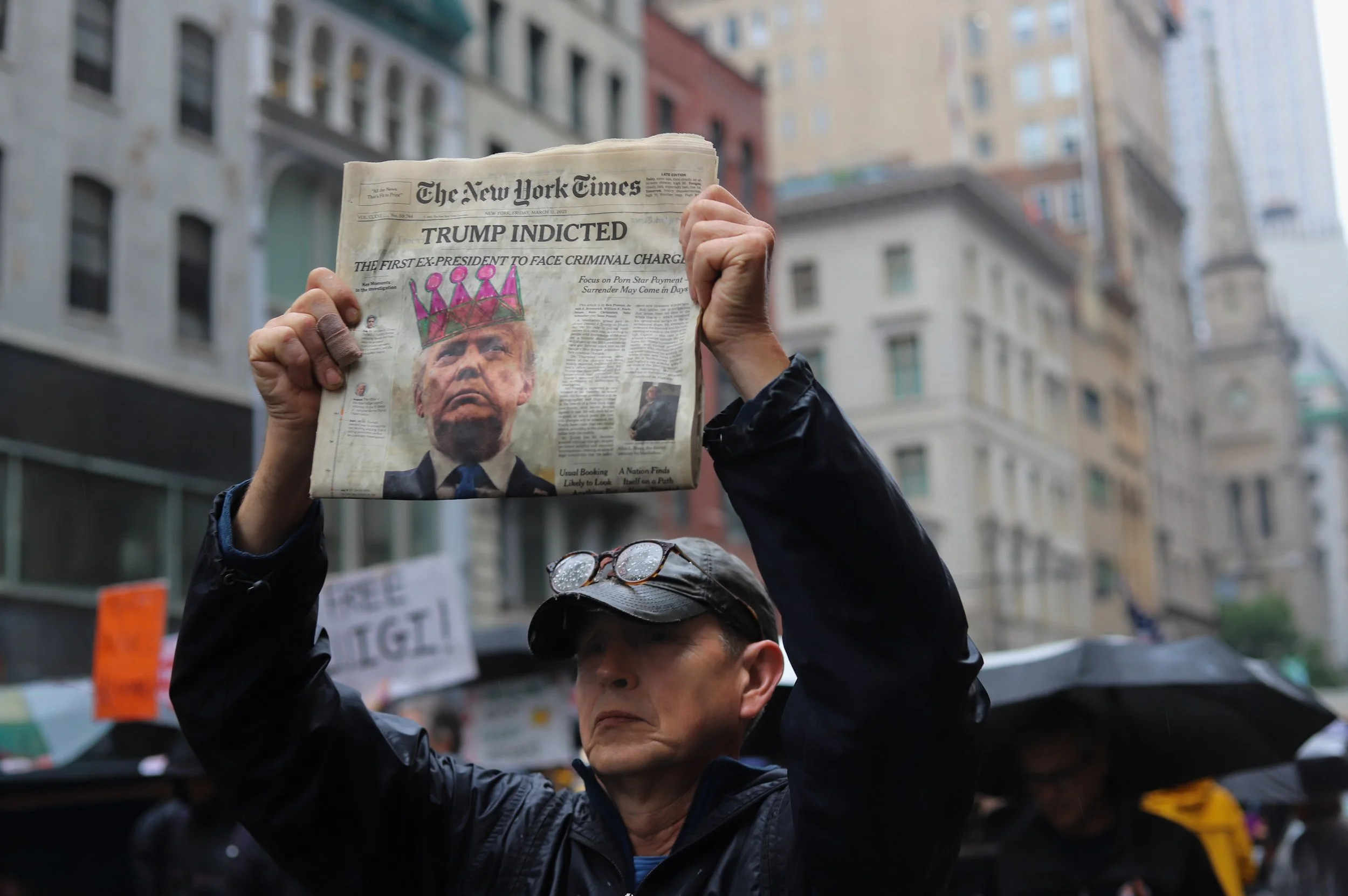 A person holding up a newspaper with the headline 'Trump Indicted' and a photo of Donald Trump wearing a crown. The setting appears to be a city street with tall buildings and other people holding umbrellas in the background.