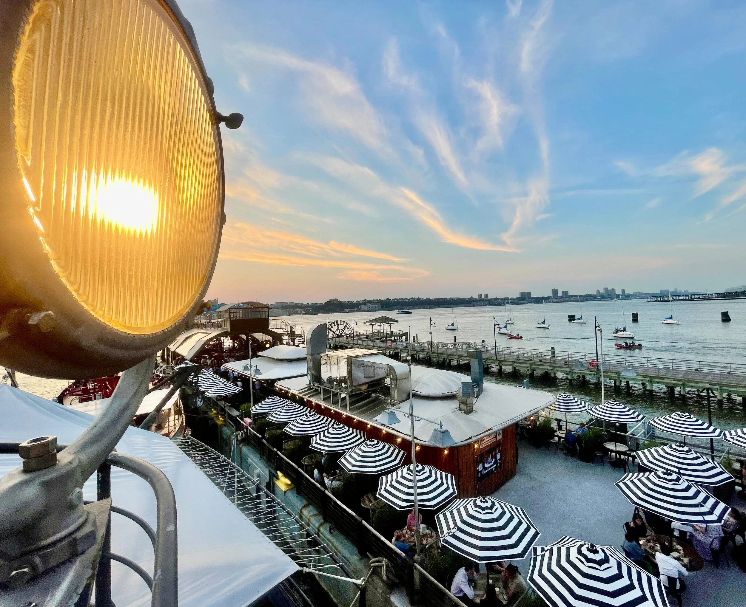 A waterfront restaurant with black and white striped umbrellas, overlooking a river with boats, during sunset with clouds in the sky.