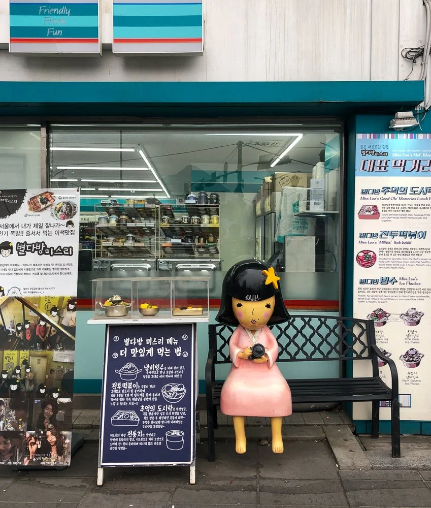 A storefront with a large doll figure sitting on a bench holding a camera. Behind the doll, there is a window with shelves of baked goods and kitchen equipment inside. To the left, there is a promotional sign with photos and text, and a blackboard si