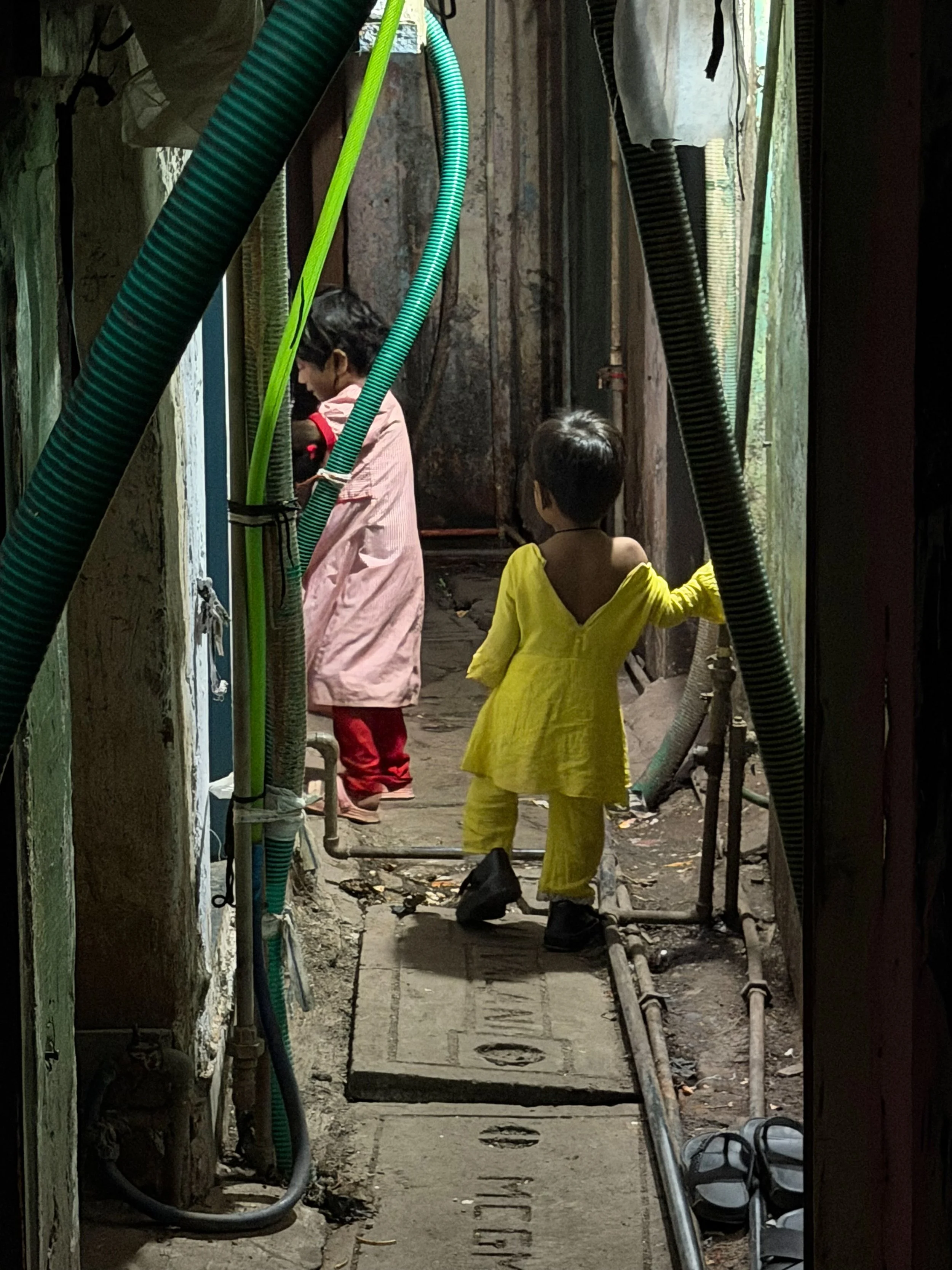 Two children in a narrow, dimly lit alleyway: a girl in a yellow outfit facing away, and a boy in a pink outfit with red pants, both surrounded by various pipes and hoses.