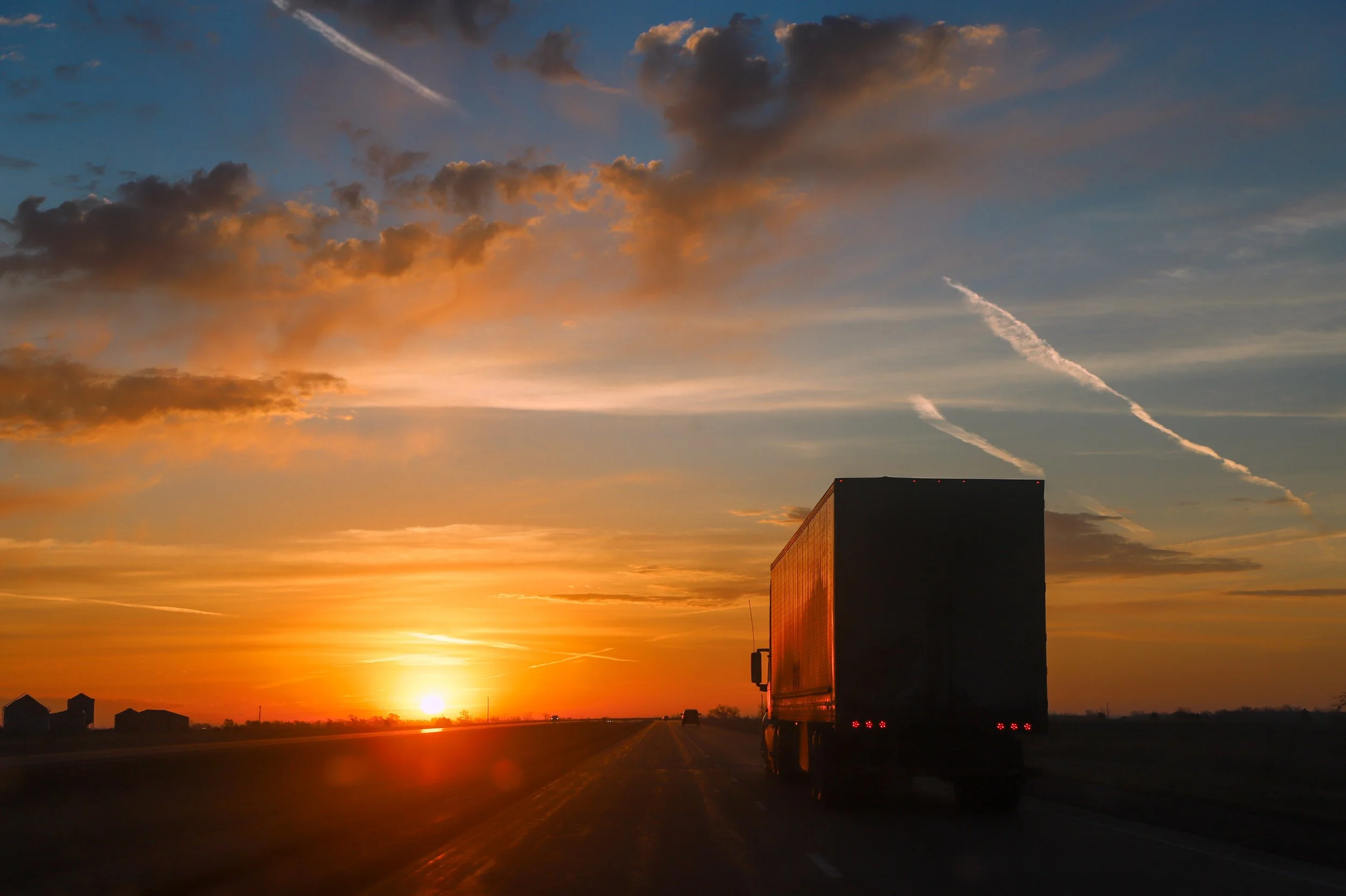 A semi-truck driving on an open road during sunset with a colorful sky filled with clouds and contrails.