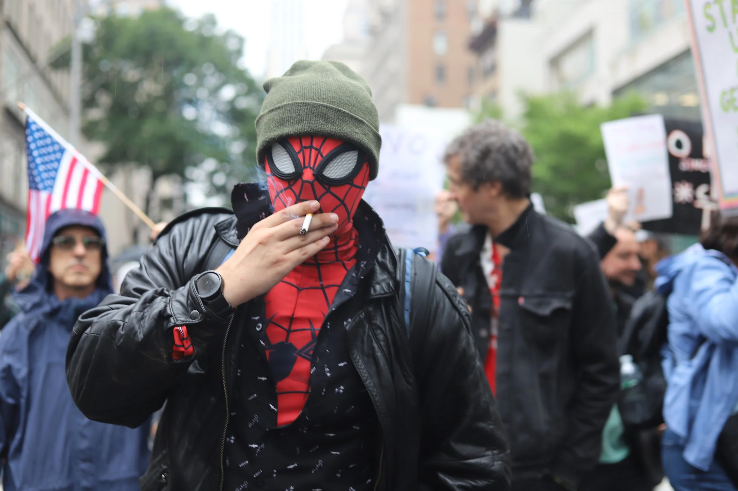 Person wearing a Spider-Man mask and a green beanie, smoking a cigarette at a protest or rally, with many other people holding signs and flags in the background.