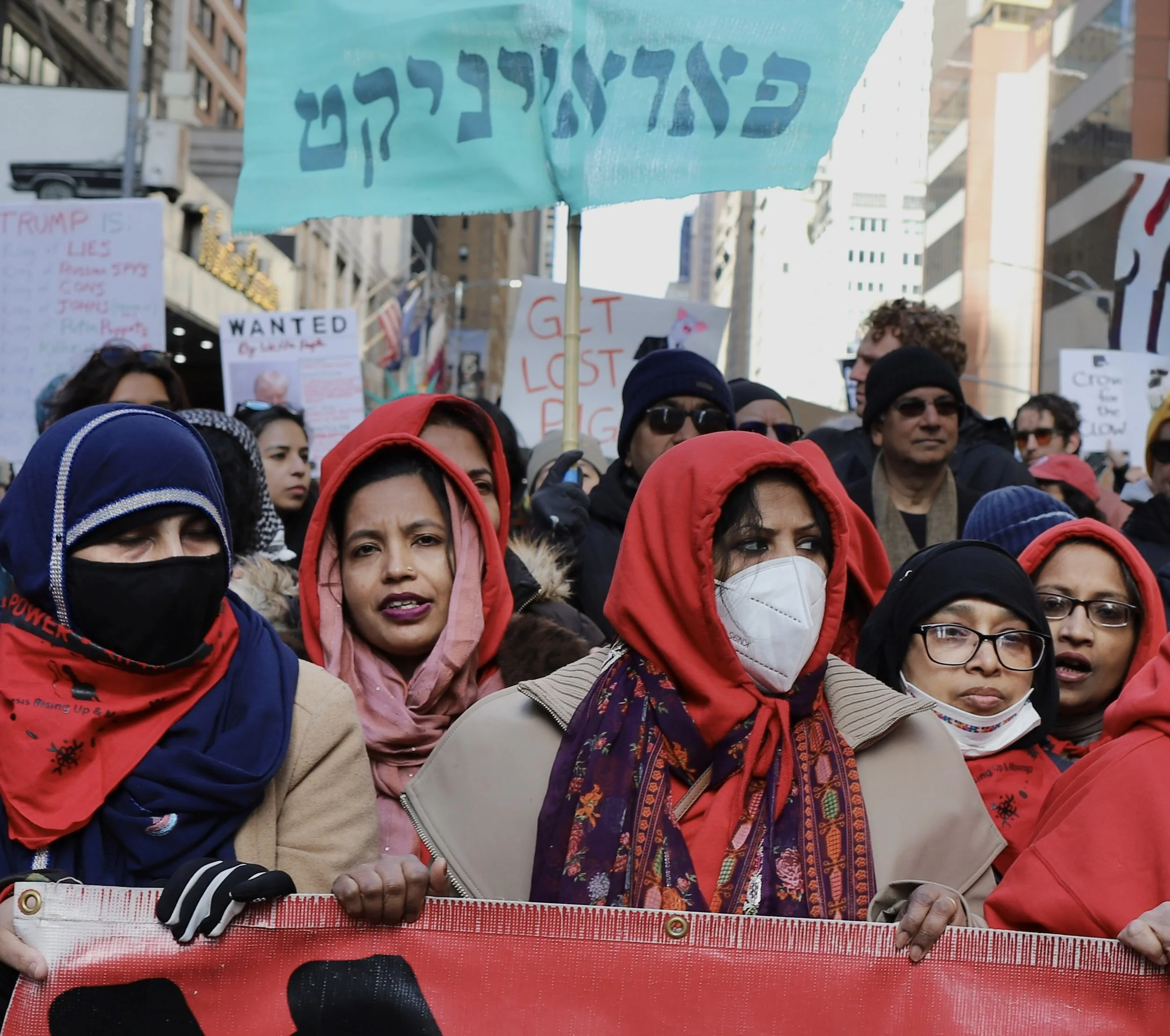 A diverse group of protesters, mostly women wearing headscarves and masks, participate in a demonstration on a city street, holding signs with messages about social and political issues.