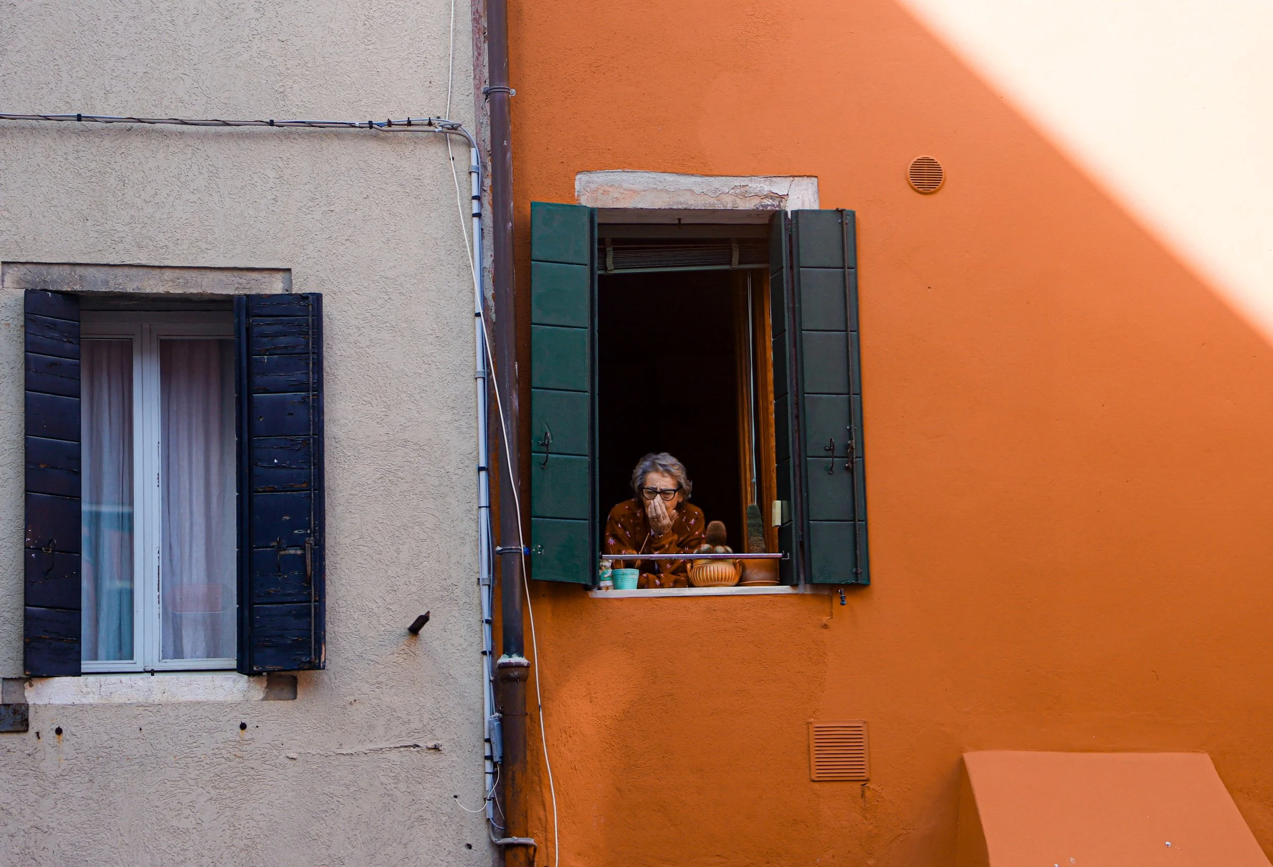 An elderly woman looking out from a small window with green shutters in an orange building, with another window with black shutters on a beige building next to it.