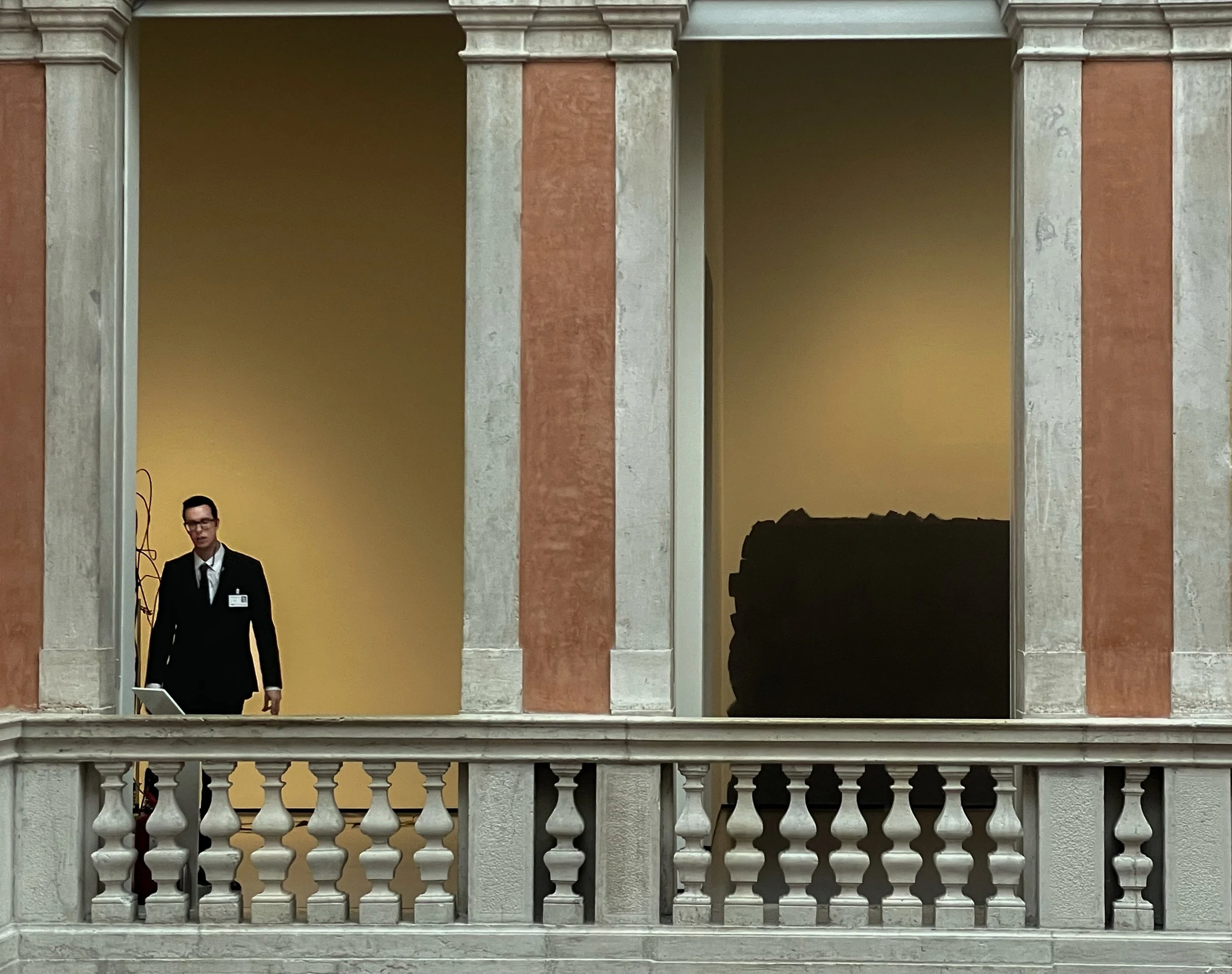 A man in a black suit with a name badge, standing on a balcony with ornate railing, in front of a yellow wall with alcoves, one containing a tall plant and the other with a black object.