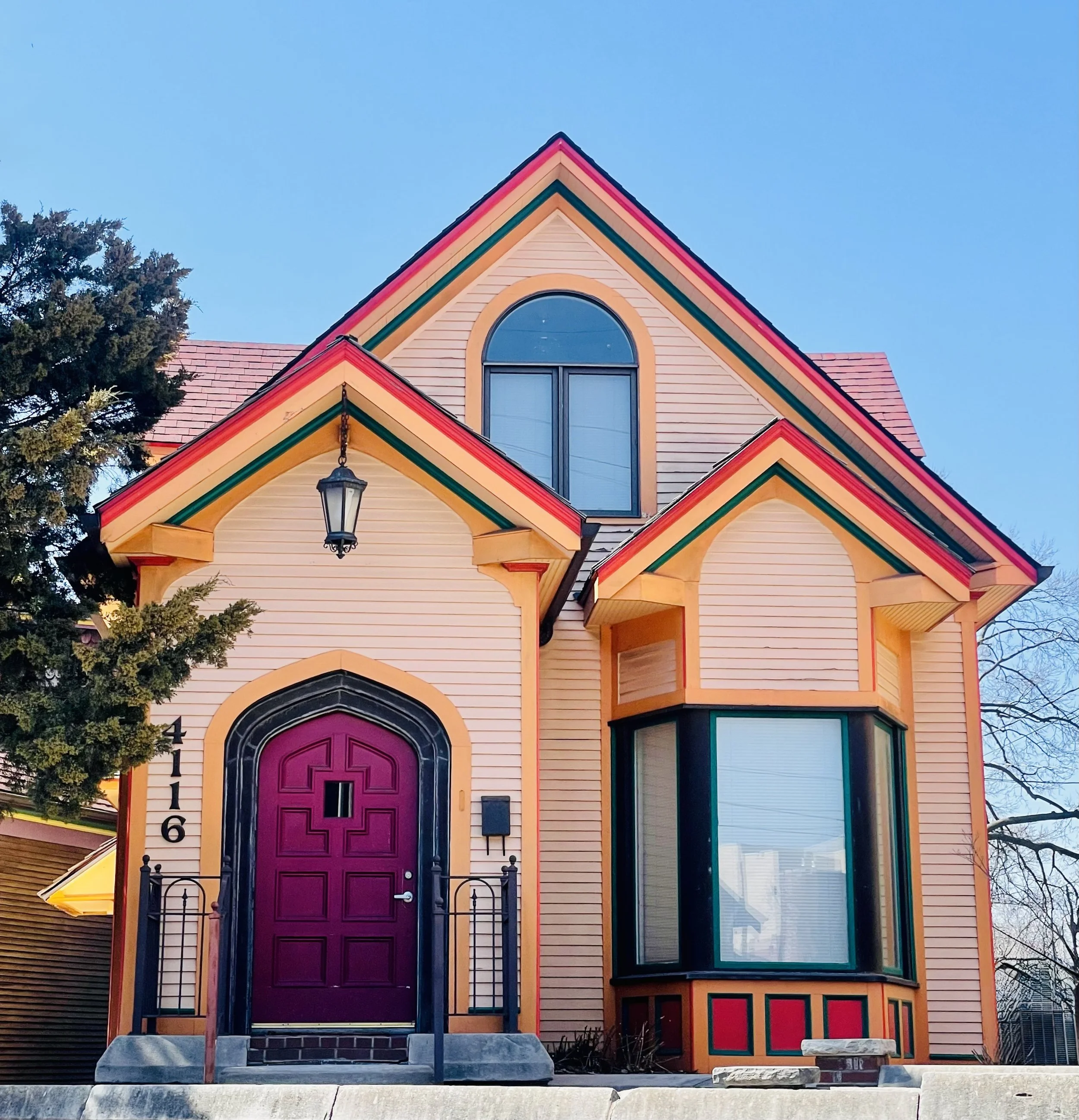 Colorful, Victorian-style house with a beige exterior, purple front door, black trim, and an arched upper window, set against a clear blue sky.
