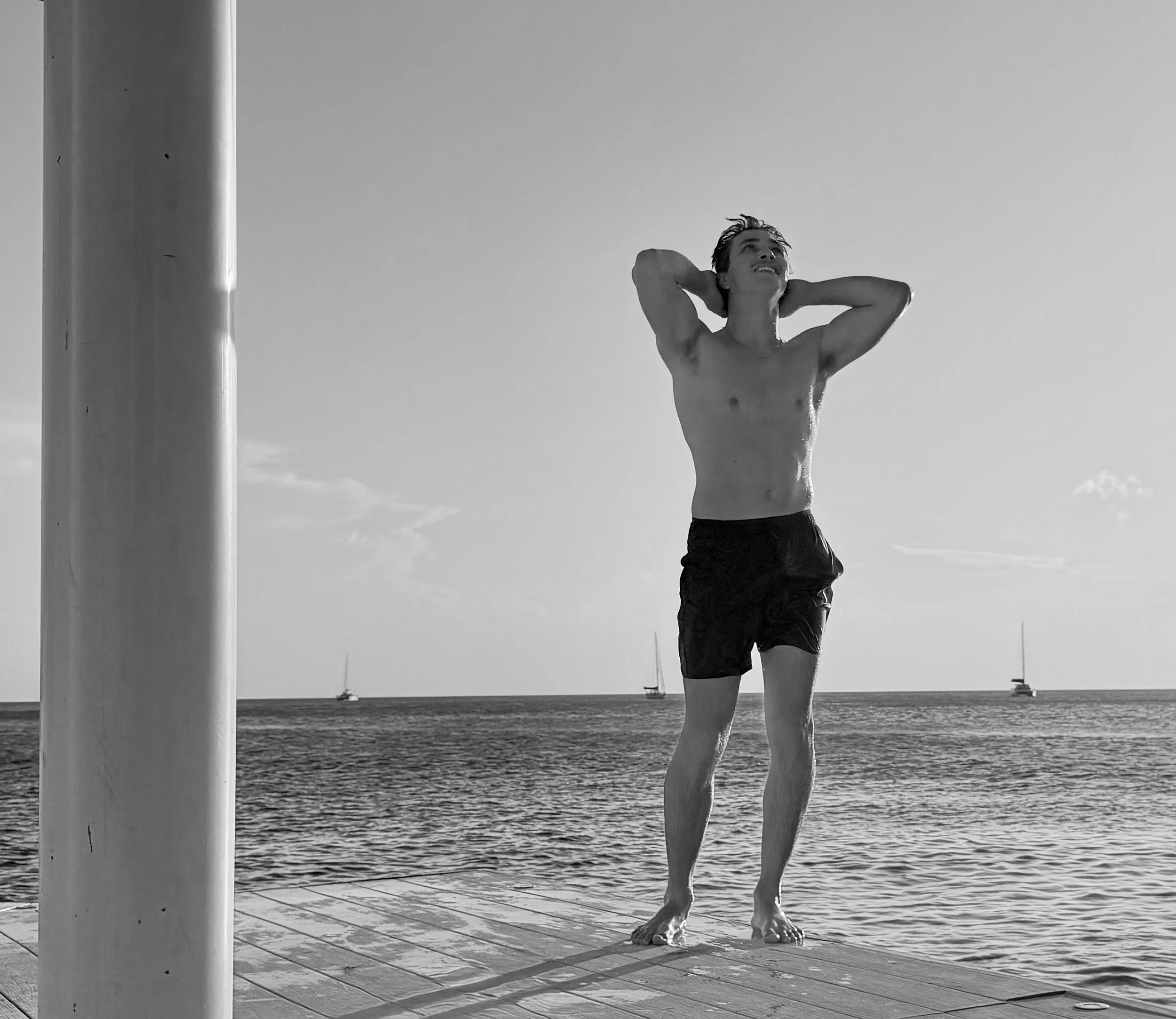 A shirtless man in black shorts standing on a dock by the water, stretching with arms behind his head, and looking up, with sailboats in the distance on a clear day.