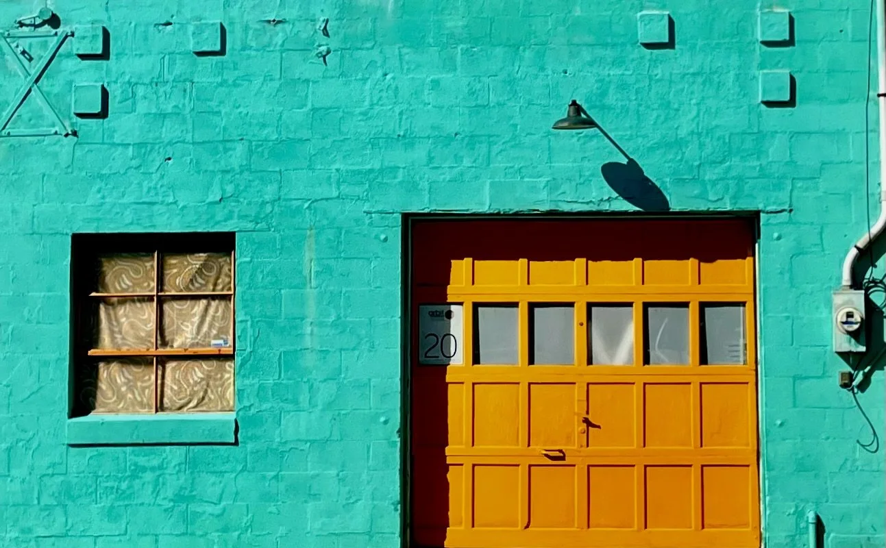 Colorful building with turquoise brick wall, yellow garage door with small windows, a small window with patterned curtains, and exterior light casting shadow