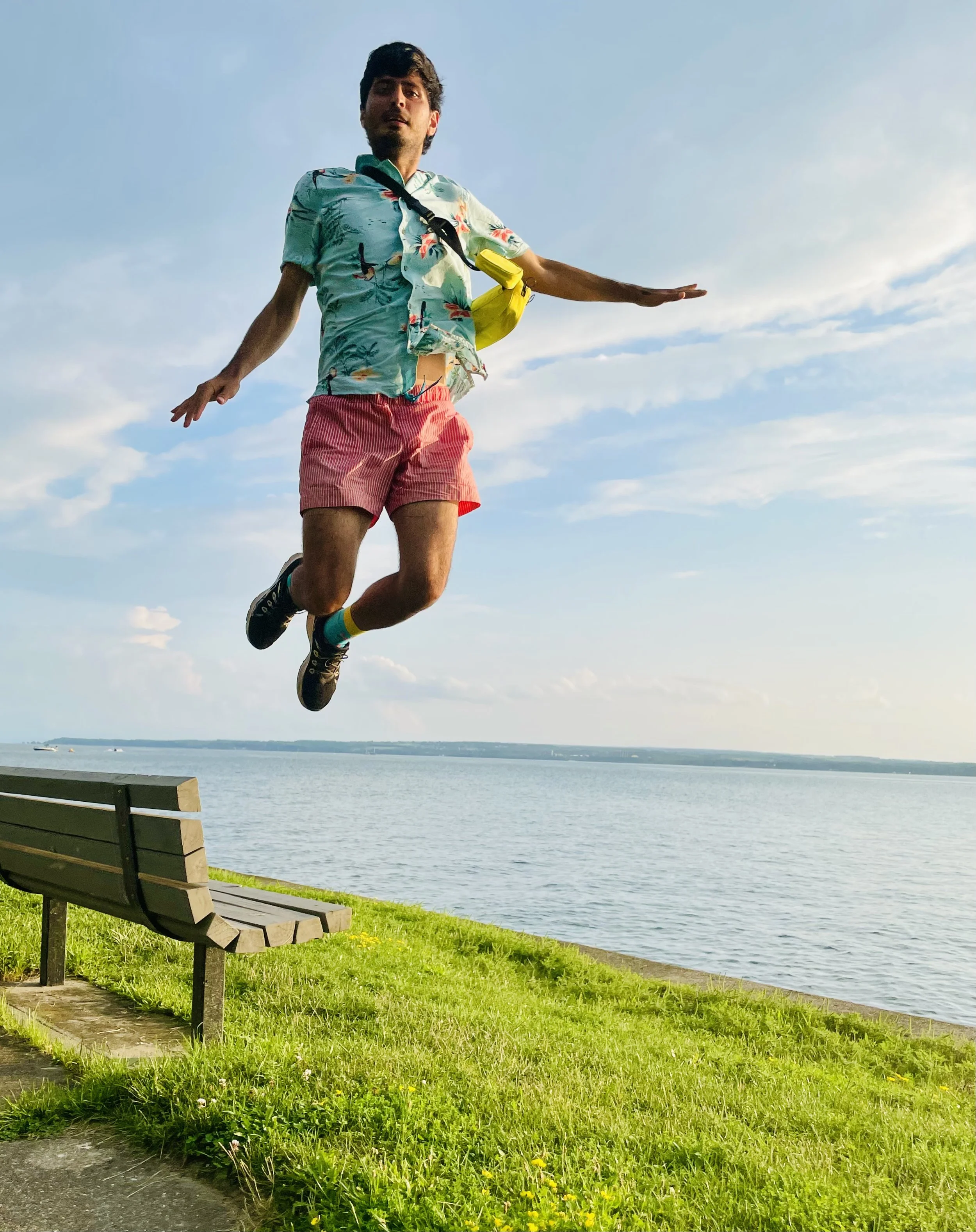 A man in casual summer clothes is jumping near a lake or ocean with a blue sky and clouds in the background.