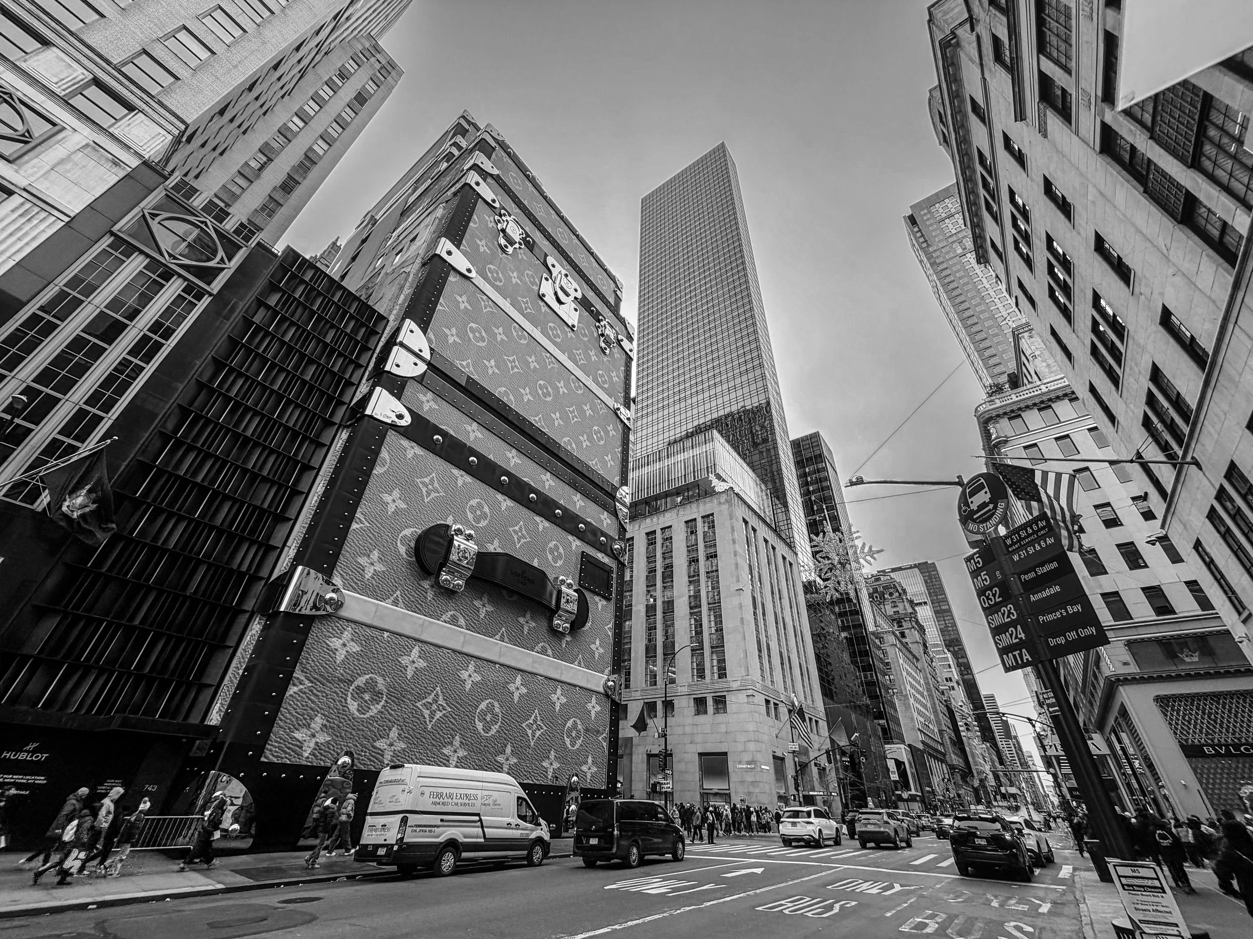 Black and white photo of a busy city street with tall buildings, a large Louis Vuitton advertisement, and people walking on the sidewalks.