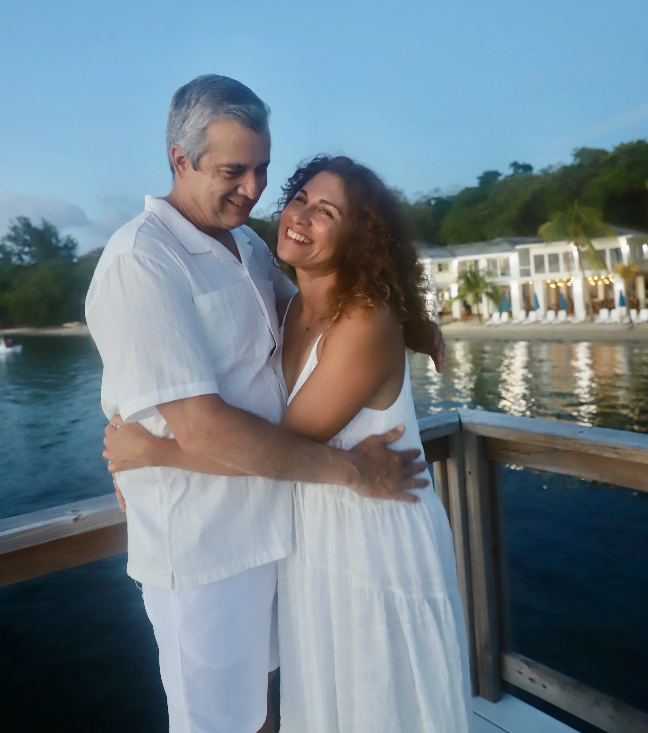 A couple hugging on a dock by the water at dusk, with a resort and tropical trees in the background.