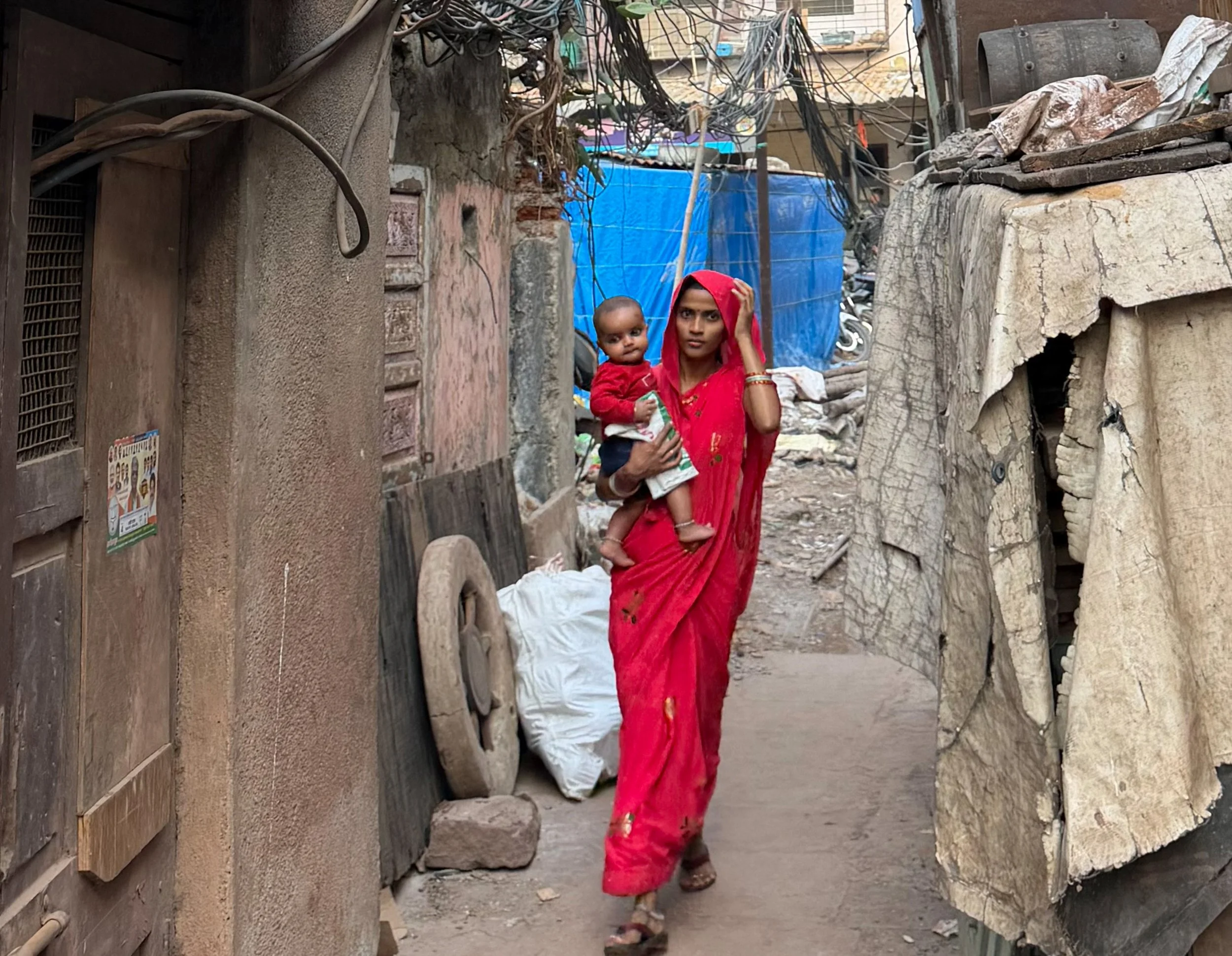 A woman wearing a red sari walking through a narrow, trash-lined alley, carrying a young child on her arm, with makeshift structures and scattered debris around.