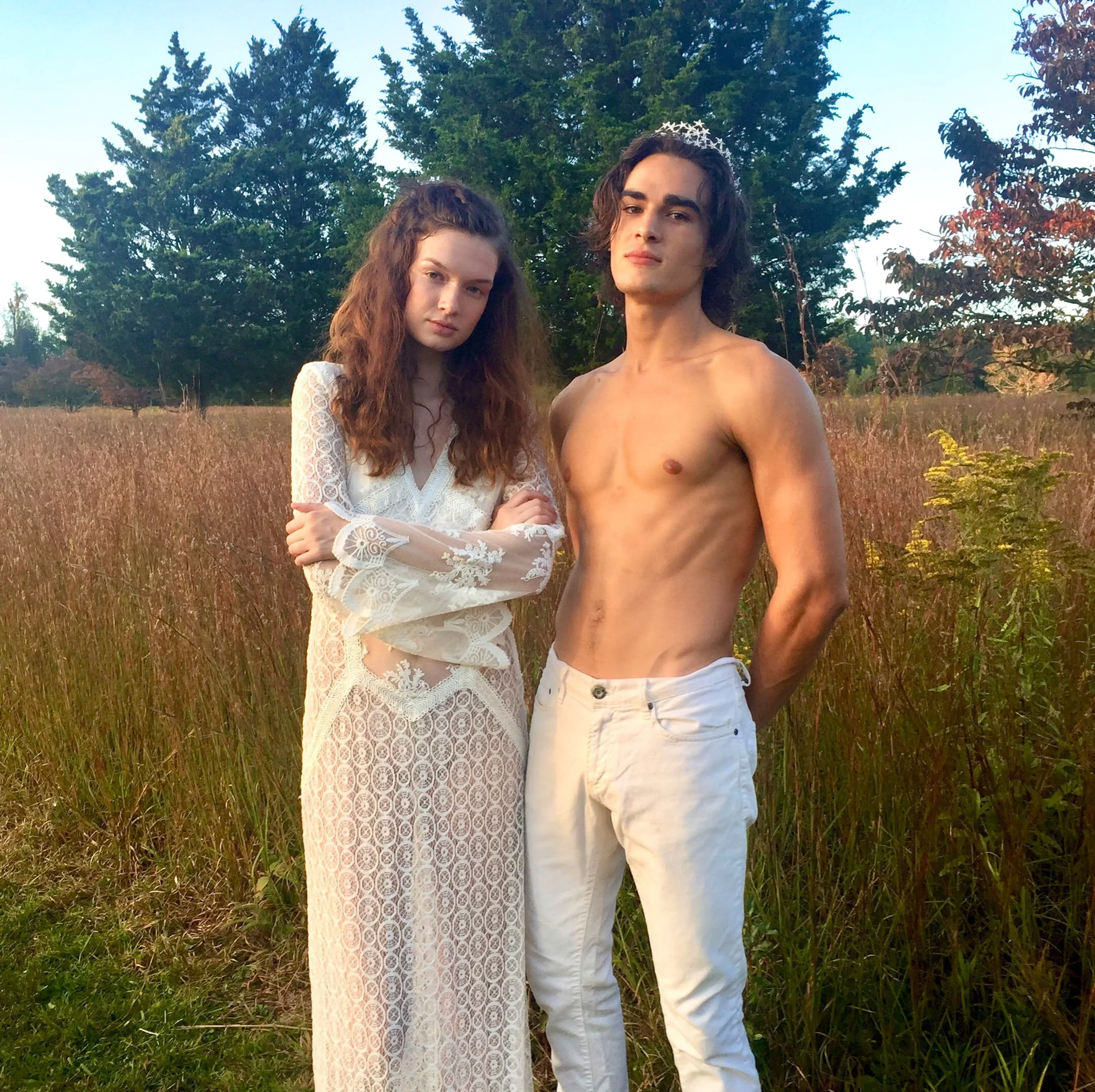 A young woman in a long white lace dress with her arms crossed and a young man shirtless wearing white pants, standing in a field of tall grass with trees in the background, during sunset.