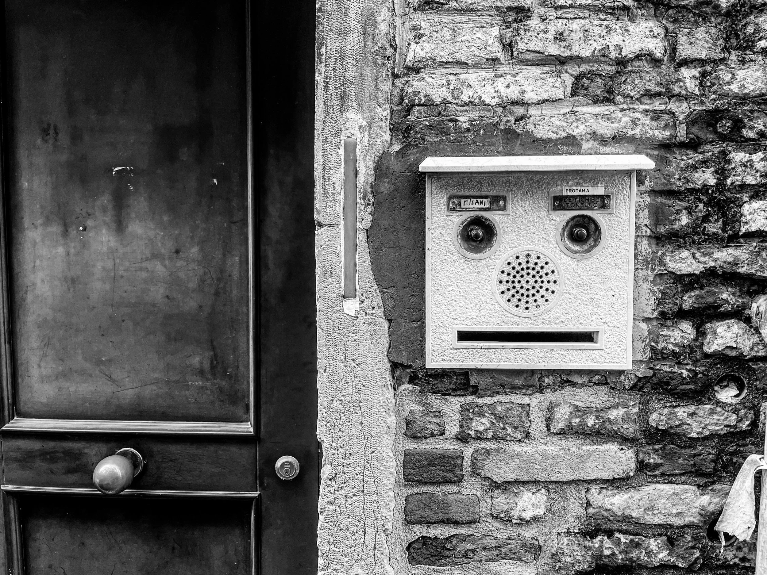 A black and white photo showing a stone wall, a metal door with a round doorknob on the left, and a humorous face-shaped mailbox with two circular openings for eyes, a round speaker grille for a nose, and a rectangular slot for a mouth on the right.