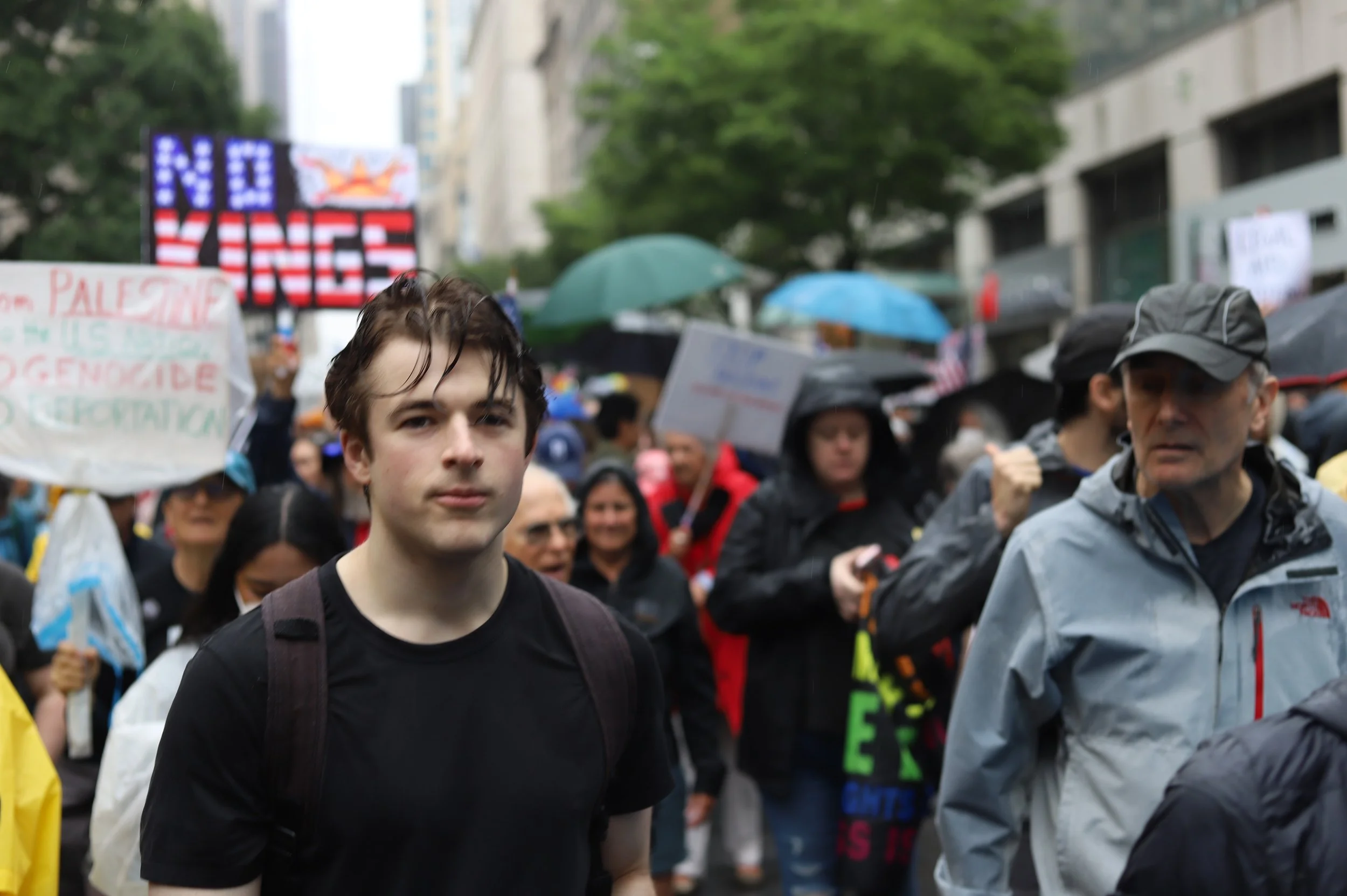 A young man in a black t-shirt at a crowded outdoor protest or rally on a rainy day, with people holding umbrellas and signs in the background.