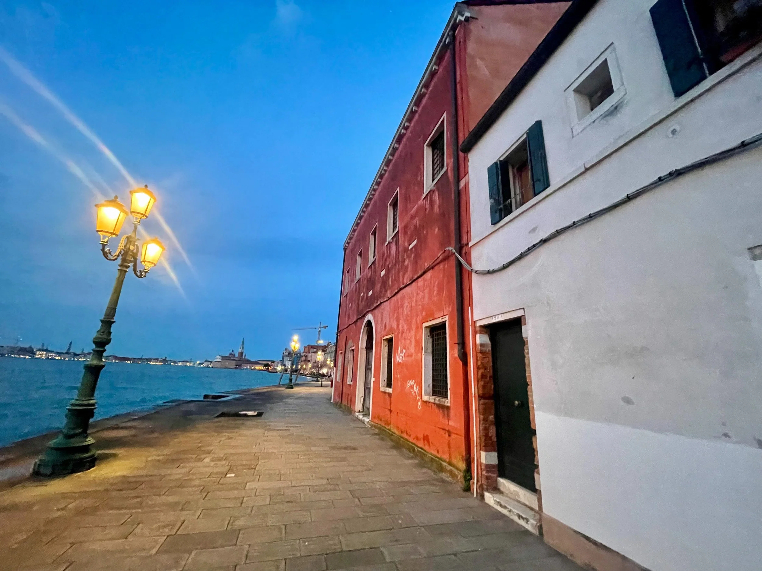 Scene of a riverside walkway with decorative street lamps, red and white buildings along the water, and distant cityscape with church steeples at dusk.