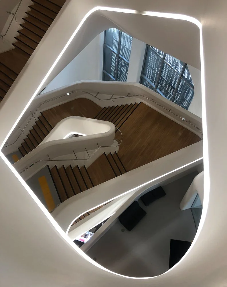 View of a modern, multi-level staircase with wooden steps, white inner walls, and large windows letting in natural light, captured from the top looking downward.
