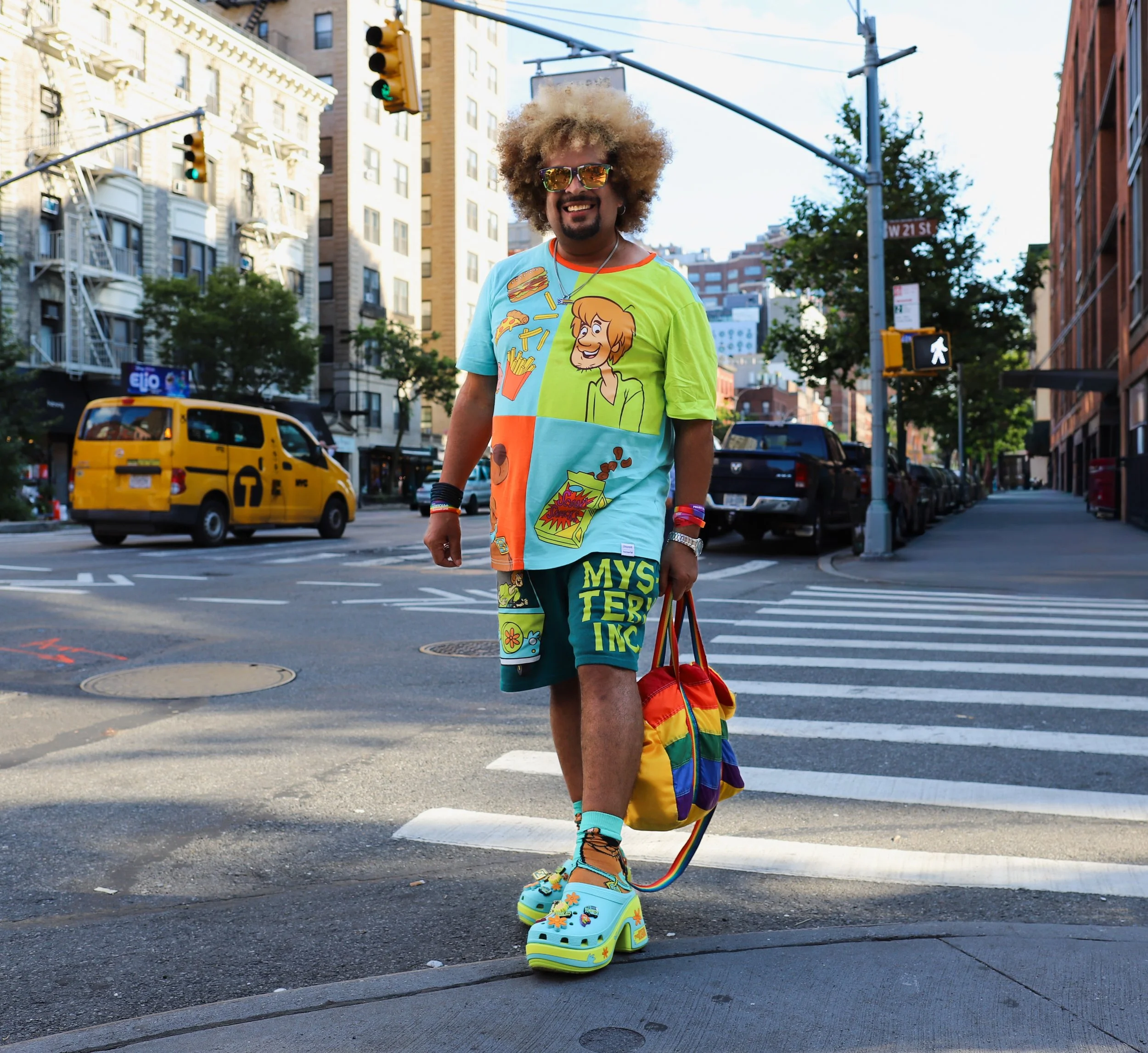 Person with curly hair and sunglasses walking across a city street wearing colorful cartoon-themed clothing and rainbow accessories, carrying a rainbow-striped bag.