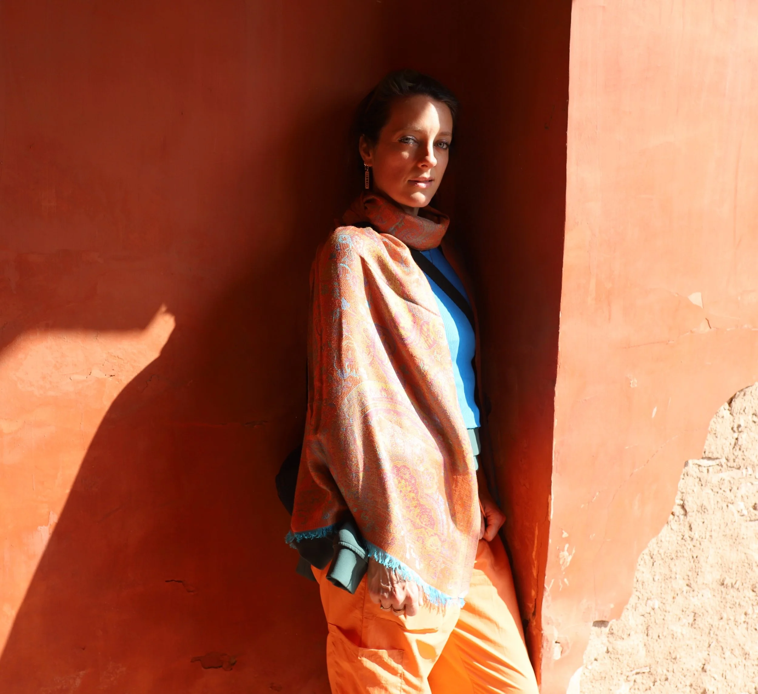 A woman leaning against an orange wall, wearing a colorful shawl, a blue top, and orange pants, with shadows cast in the sunlight.