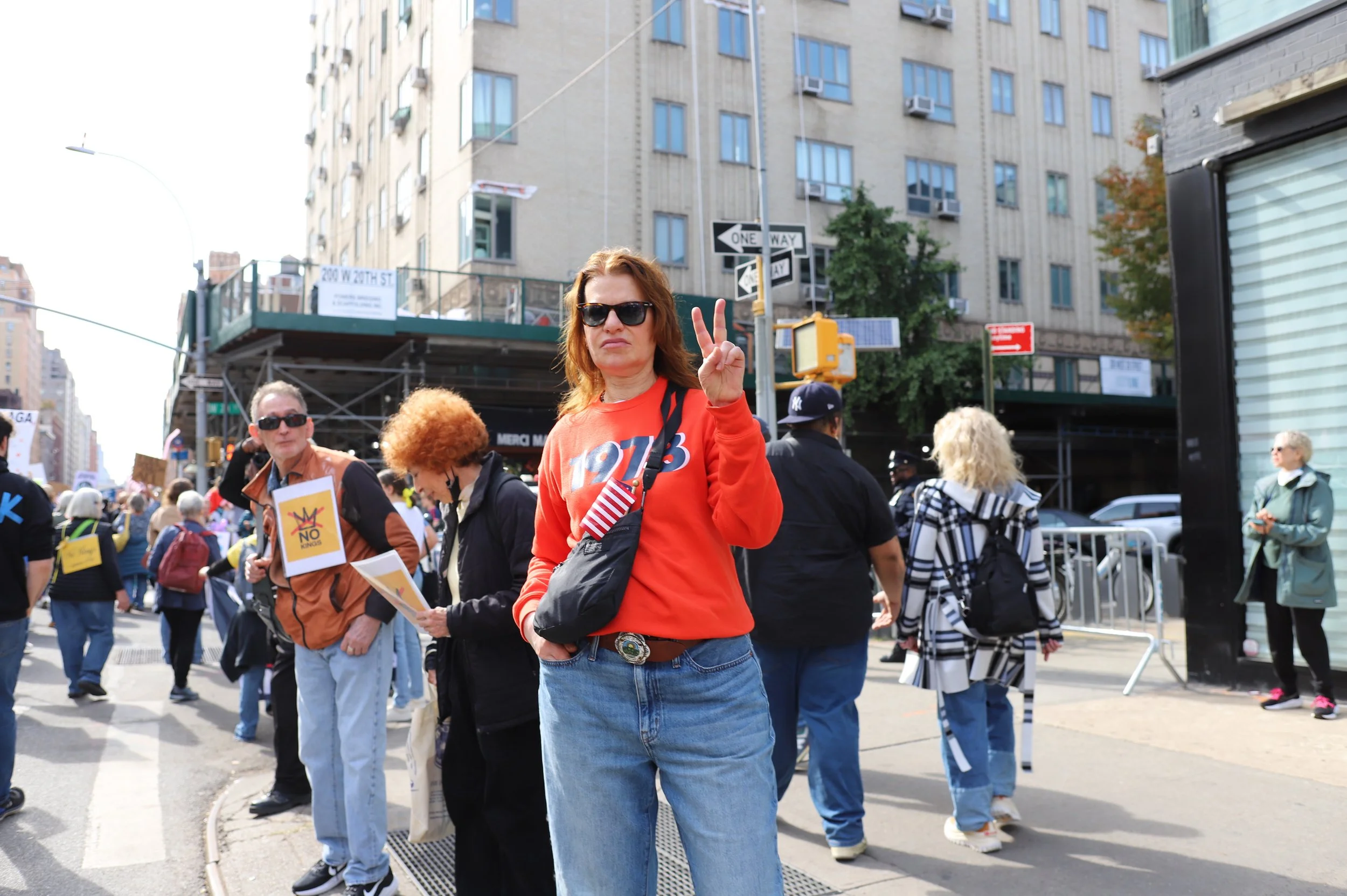 A woman with sunglasses and a red sweatshirt making a peace sign at a crowded protest or march on city streets.