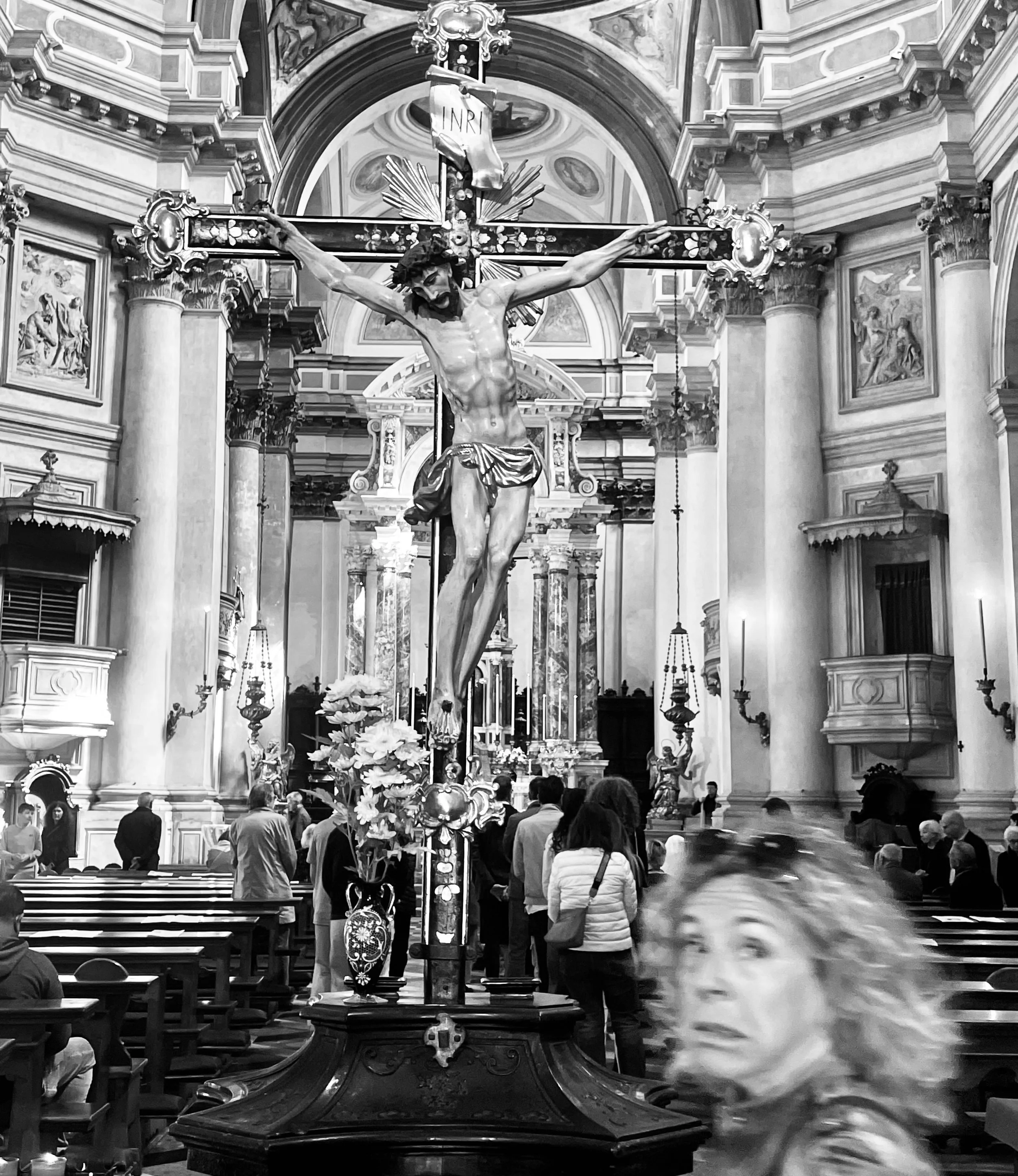 A large religious statue of Jesus Christ crucified on the cross inside a church, surrounded by ornate architectural details and people praying.