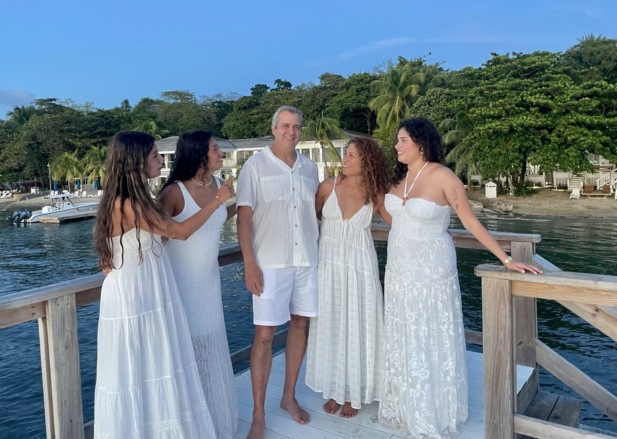 A group of five people standing on a wooden dock by the water, dressed in white summer clothing, with trees and houses in the background during daytime.