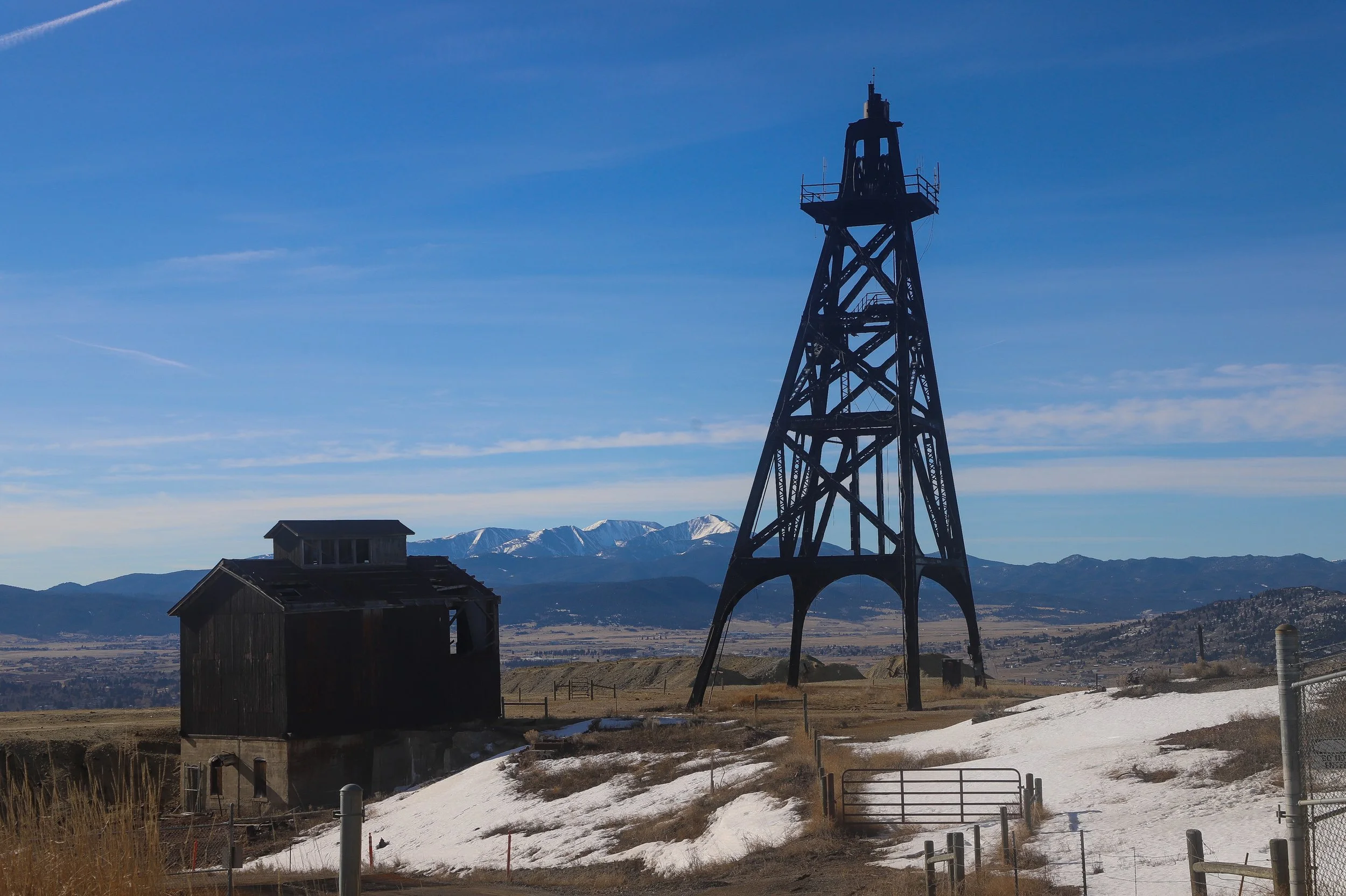 An old wooden barn next to a tall metal oil derrick with snow patches on the ground and mountains in the background under a blue sky.